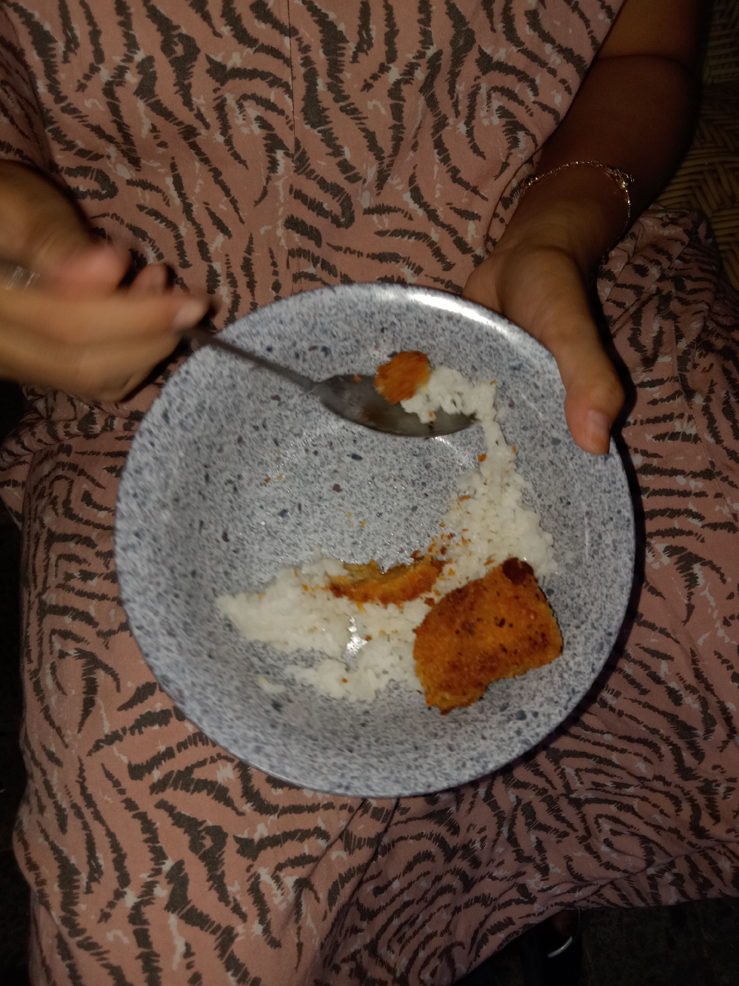 In Jumput, Indonesia, a close-up shot shows a person holding a spoon over a bowl of rice with a piece of fried chicken. The person is wearing a pink dress with a dark, abstract pattern. The bowl is gray with a speckled texture. The fried chicken appears golden brown and is placed on top of the white rice. A silver spoon is positioned above the food, with a small portion of rice on its tines. The lighting suggests an indoor setting, possibly at night.