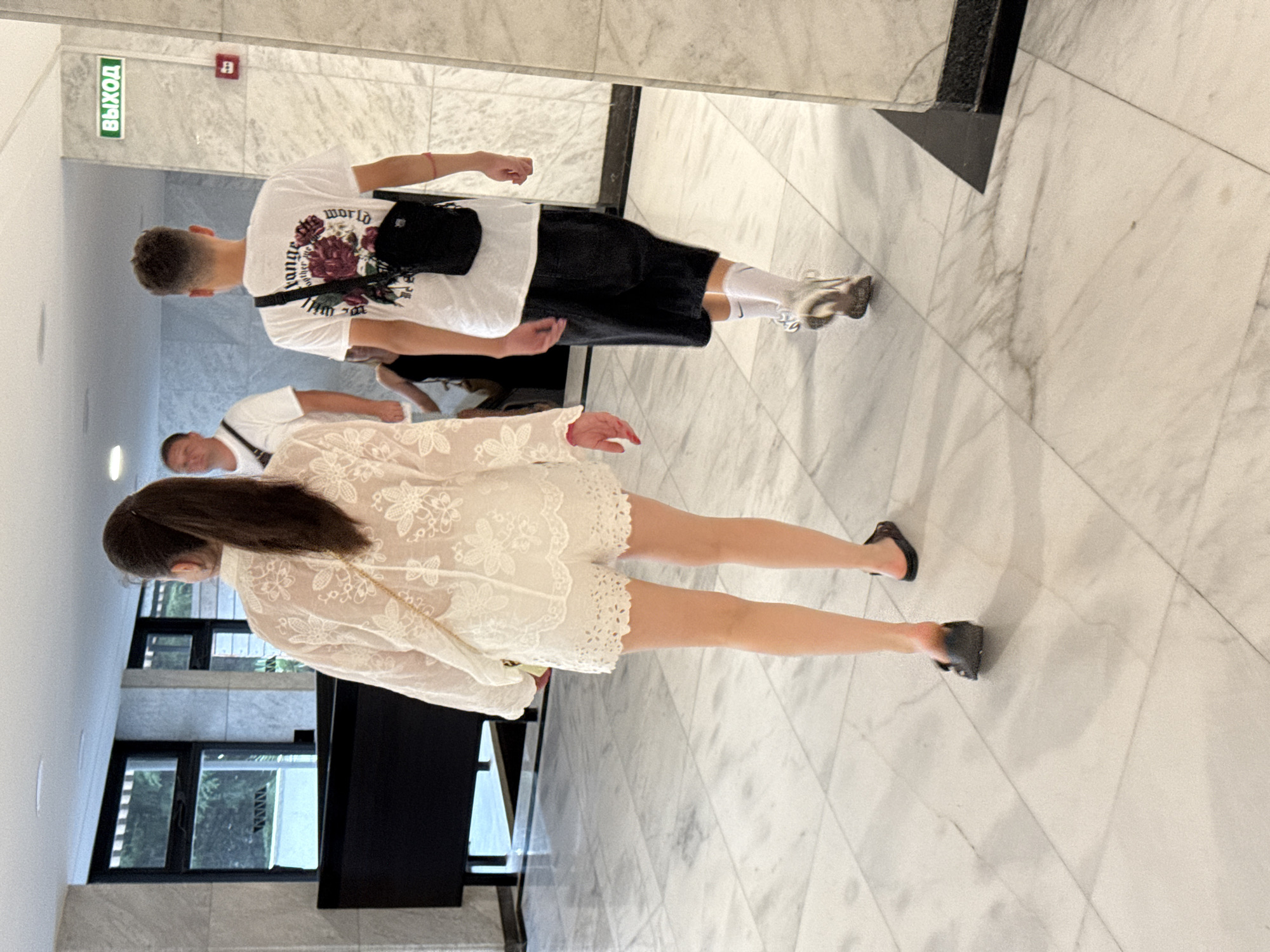 The image shows three individuals walking away from the camera in an indoor, well-lit hallway with marble floors. The person in the foreground on the left is a woman with dark hair pulled back in a ponytail, wearing a white lace top and white lace shorts, and dark sandals. She is carrying a small light-colored purse over her shoulder. To the right of her, a young man is walking, wearing a white t-shirt with a floral graphic on the back, black shorts, white socks, and athletic shoes. He is wearing a black crossbody bag. Behind them and to the left, another man is walking, wearing a white t-shirt and a dark crossbody bag. A green sign with the word "ВЫХОД" (Exit in Russian) is visible on the right wall, along with a red fire alarm. The environment appears to be a hotel lobby or a similar public building in Yalta, Ukraine, given the Russian signage.