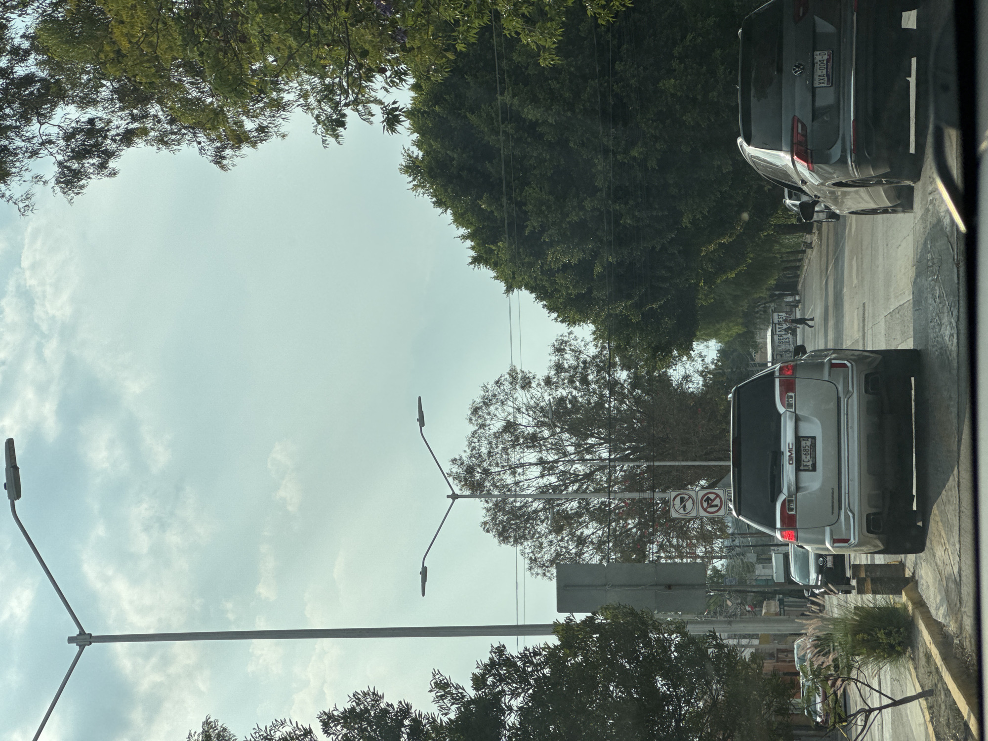 The image shows a street scene in Puebla, Mexico, likely taken from inside a vehicle. A line of cars is visible on the right side of the road, with a silver GMC SUV in the foreground and a dark Volkswagen behind it. The SUV has the license plate JFC-685-A, and the Volkswagen has the plate XXA-004-D. Streetlights with a distinctive Y-shape are present, and traffic signs are also visible. The road is bordered by sidewalks and greenery, including trees that overhang the scene. The sky is overcast with patchy clouds.