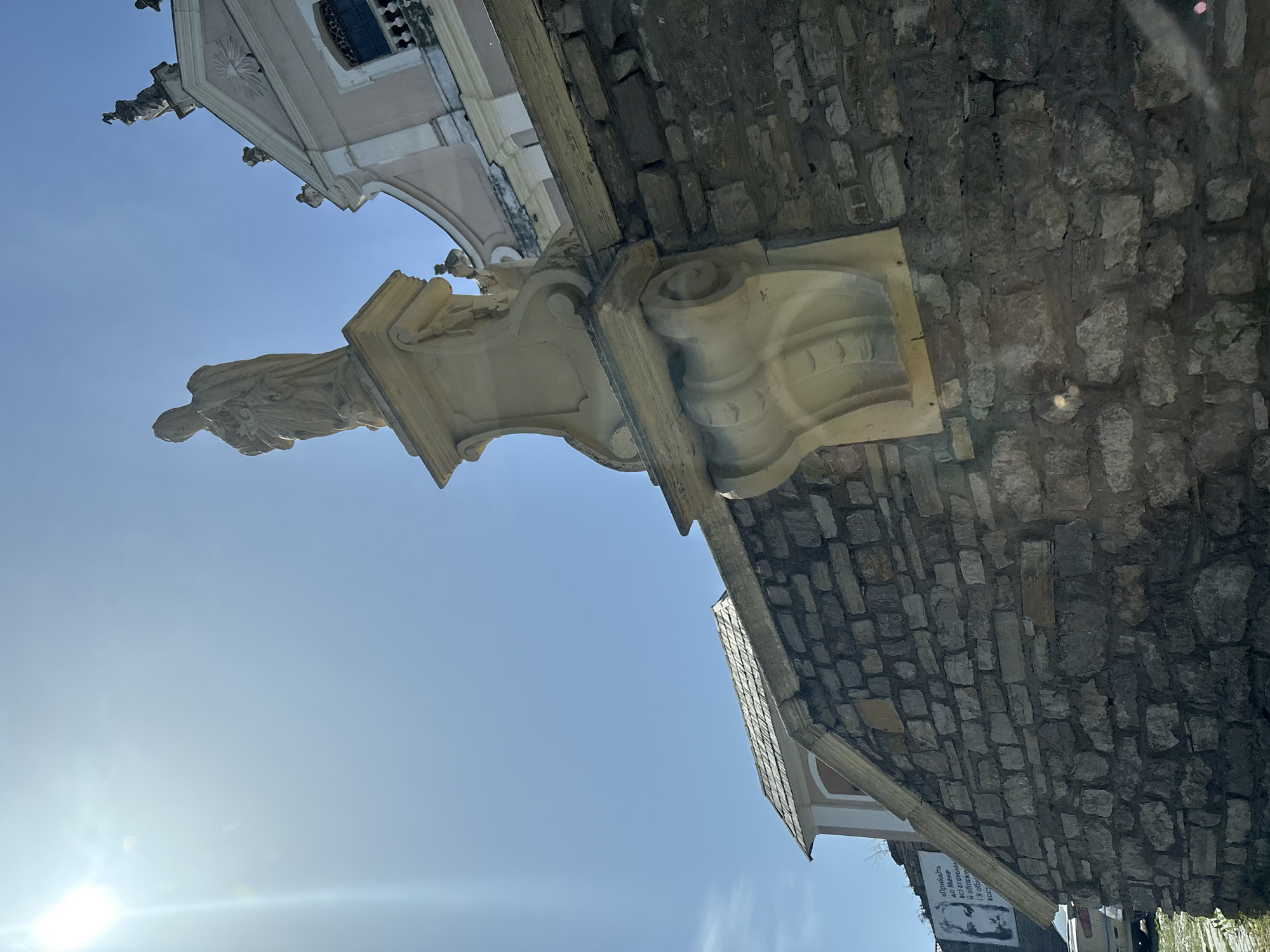 A statue of a person stands atop a stone pedestal against a bright blue sky. The statue is made of stone and appears to be draped in robes. To the right of the statue, a pink and white building with ornate architectural details is visible. In the foreground, a rough stone wall runs diagonally across the frame. The sun is shining brightly, casting a lens flare in the upper left corner. A street sign with Ukrainian text is visible on the stone wall in the lower left corner.