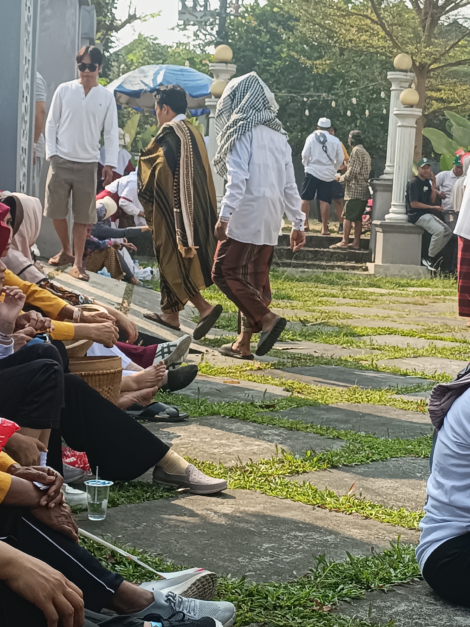 In a city scene in Sleman, Indonesia, a group of people are gathered outdoors, possibly at a religious or community event. Sunlight filters through trees, casting shadows on a paved walkway with patches of grass.
In the foreground, several individuals are seated on the ground. A person in a yellow shirt and black pants is visible, with hands clasped. To their left, another person in black pants and a yellow top sits with their hands resting on their lap. A cup of water with a straw is placed beside them. Further to the right, a person in a white shirt and black pants is seated, with their legs crossed and wearing a light-colored slip-on shoe. Another person in a white shirt and black pants is also seated to the far right of the frame.
In the midground, a man in a white long-sleeved shirt and beige shorts, wearing sunglasses, stands with his hands in his pockets. He is looking towards the right. To his right, a man in a traditional brown and gold garment with intricate patterns walks away from the camera. He is wearing a checkered headscarf and dark sandals. Another man in a white long-sleeved shirt and brown pants is walking past him. In the background, other people are visible, some standing and some seated, and a blue and white umbrella is set up. Decorative lights are strung across trees.
The environment suggests a warm, sunny day, with the people dressed in light clothing suitable for the Indonesian climate. The paved walkway and grassy areas indicate an outdoor public space. The presence of traditional attire suggests a cultural or religious gathering.