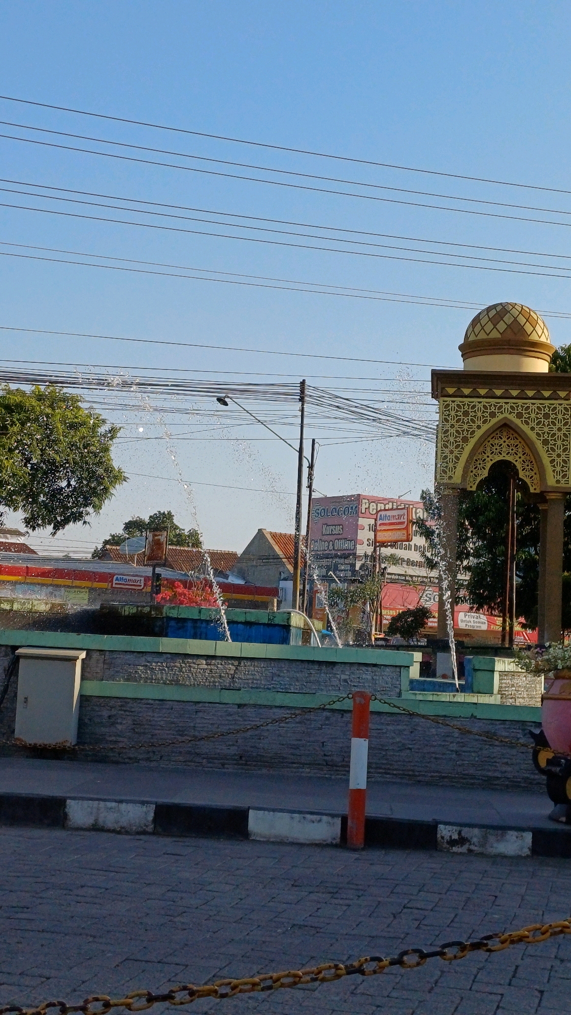 This image captures a street scene in Jonggrangan, Indonesia, featuring a public fountain with water spraying upwards. To the right, an ornate, domed structure with Islamic architectural elements stands prominently. In the background, a tree is visible on the left, and various buildings, including an Alfamart convenience store, are present. Overhead, a network of power lines crisscrosses the clear blue sky. The foreground shows a paved sidewalk with a decorative chain fence and a bollard painted with orange and white stripes. The overall atmosphere suggests a sunny day in a populated urban area.