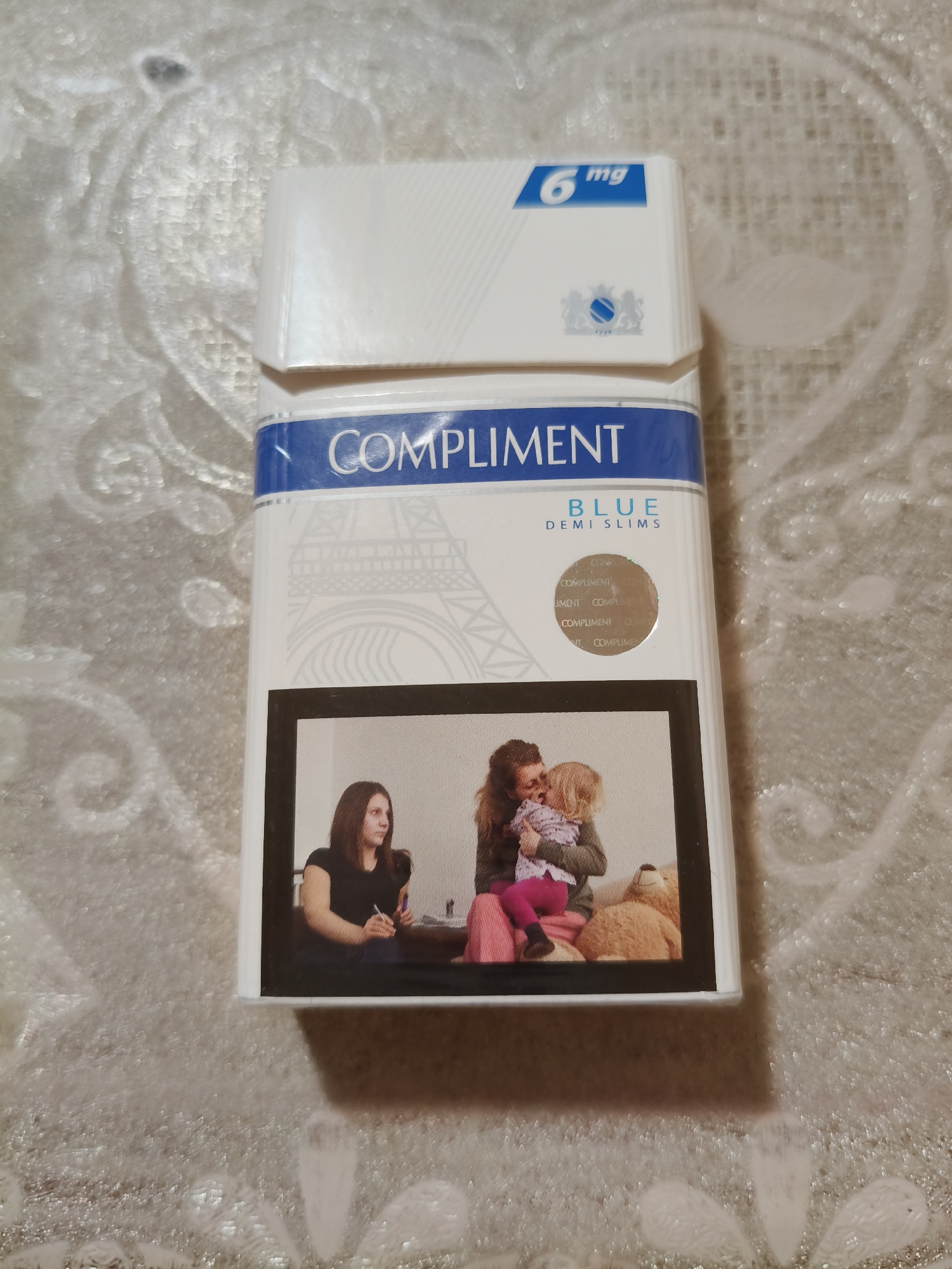 An overhead shot captures a pack of "Compliment Blue Demi Slims" cigarettes resting on a textured white surface. The pack is predominantly white with a blue band across the top, featuring the brand name in white capital letters. Below the brand name, a graphic of the Eiffel Tower is subtly imprinted in light grey. The bottom portion of the pack displays a photograph framed in black, depicting two women and a child. The woman on the left, wearing a black top, is looking to her right, holding a pen. To her right, another woman in a green sweater and pink pants holds a young child, who is reaching out. A teddy bear sits to the right of the women and child. The image does not contain any information that would suggest its location in Chornobaivka, Ukraine, or any other specific city.