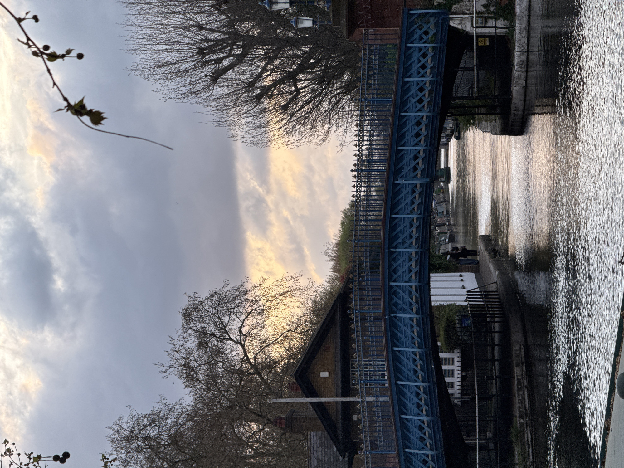 The image depicts a blue metal bridge spanning a waterway in London, United Kingdom. The canal reflects sunlight. Adjacent to the bridge is a building with a dark roof and light brick walls. Bare trees frame the scene against a cloudy sky with patches of light. Pedestrians are visible on the towpath.