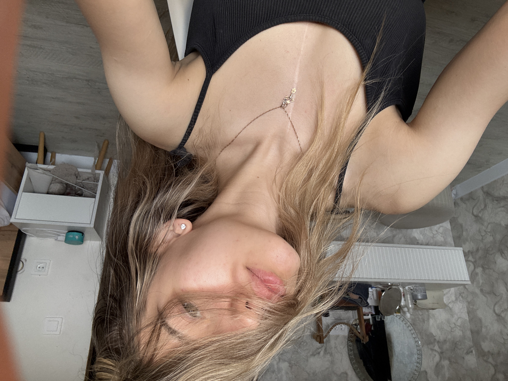A selfie, taken from above, shows a young woman with light blonde hair. She is wearing a black tank top and a delicate necklace with a pendant that rests on her upper chest, where a faint vertical scar is visible. The image is rotated, with the woman's face appearing sideways in the frame. Her left eye is visible, and part of her hair covers the right side of her face. The background includes a white storage unit with items inside, an electrical outlet, and a floor with a gray marble-like pattern. A lamp is also visible in the background. There is no information in the image to suggest that this was taken in Noviy, Russia.