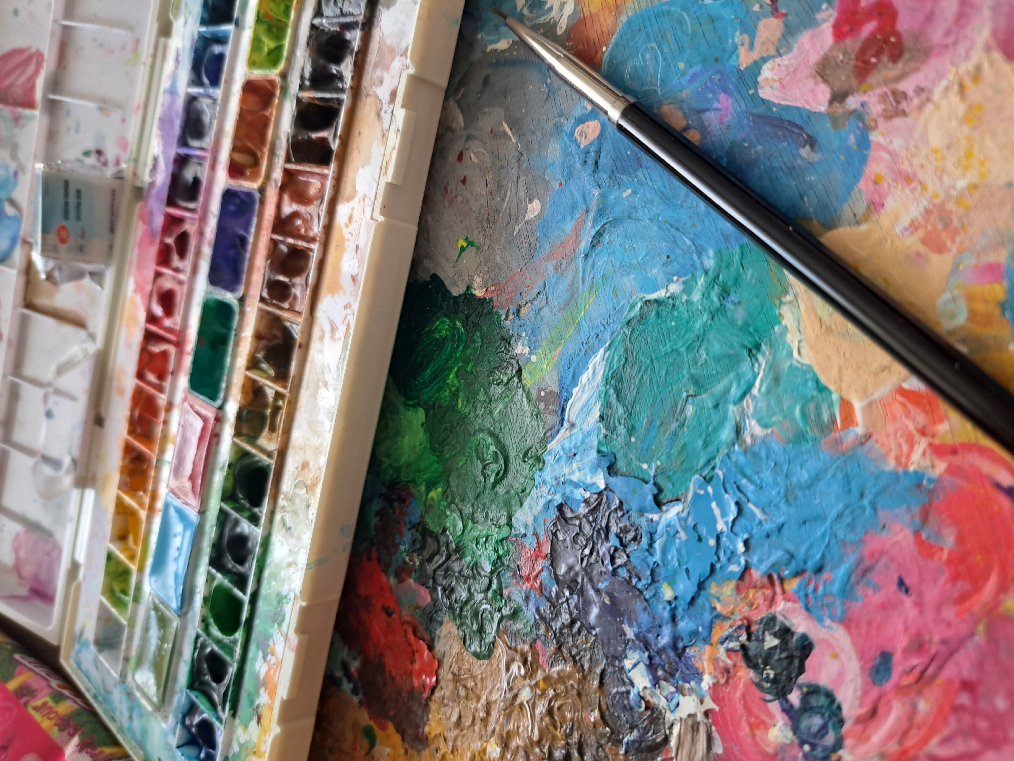 The image displays a close-up view of art supplies, specifically a watercolor paint palette and a painter's palette with dried paint. The watercolor palette is filled with numerous individual pans of paint in various colors, including blues, greens, reds, oranges, purples, and black. Some pans appear nearly full, while others are visibly used. Next to the watercolor palette is a larger, well-used painter's palette, a flat surface heavily encrusted with layers of dried paint in a vibrant, abstract pattern of blues, greens, pinks, reds, and browns. A black-handled paintbrush with a silver ferrule rests on top of this painted surface, its tip touching some of the dried paint. The overall impression is one of an active art studio or workspace. There is no indication of people, specific time of day, weather, or strong emotional cues, beyond the implication of creative activity. The location context of Popovo, Bulgaria, does not provide any visual cues within the image itself. A small piece of packaging with Cyrillic text is visible on the left side of the watercolor palette, though the text is largely illegible.