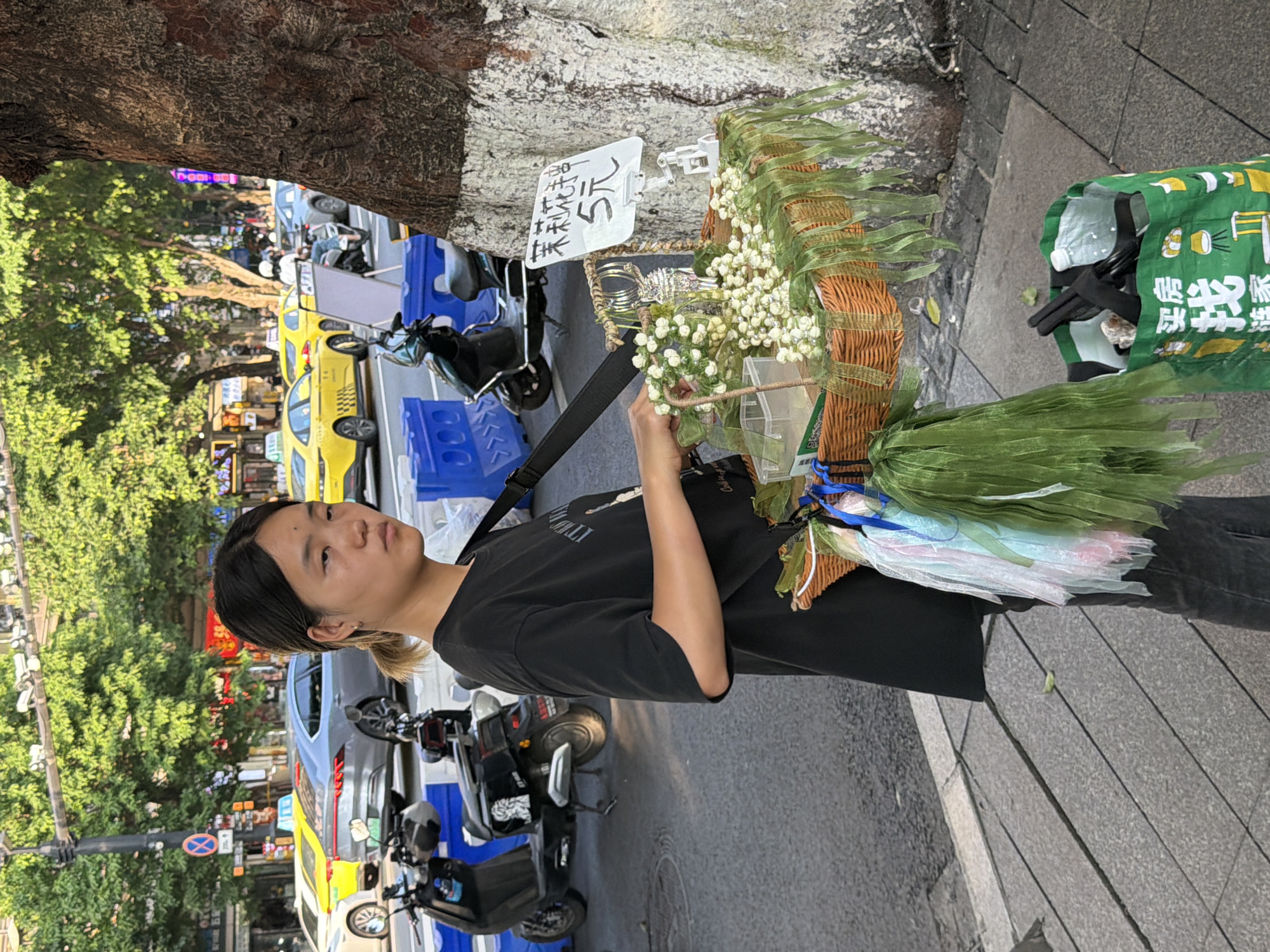 A young Asian female, appearing to be in her late teens or early twenties, stands on a sidewalk in Yuzhong District, China, selling small white flowers from a woven basket. She is wearing a black t-shirt and dark pants. The basket, which hangs from her shoulder by a dark strap, is decorated with green ribbons and colorful fabric streamers. A sign displaying Chinese characters and the number "5元" (5 Yuan) is attached to a white stand next to the basket. In the background, a tree trunk with white markings is visible on the right. The sidewalk is made of gray concrete slabs. Behind the sidewalk, a street with yellow taxis and other vehicles is present, along with more trees and buildings. Two dark-colored scooters are parked on the street. A green reusable shopping bag filled with various items sits on the sidewalk at the lower right of the frame. The lighting suggests it is daytime, likely late afternoon, with sunlight casting shadows.