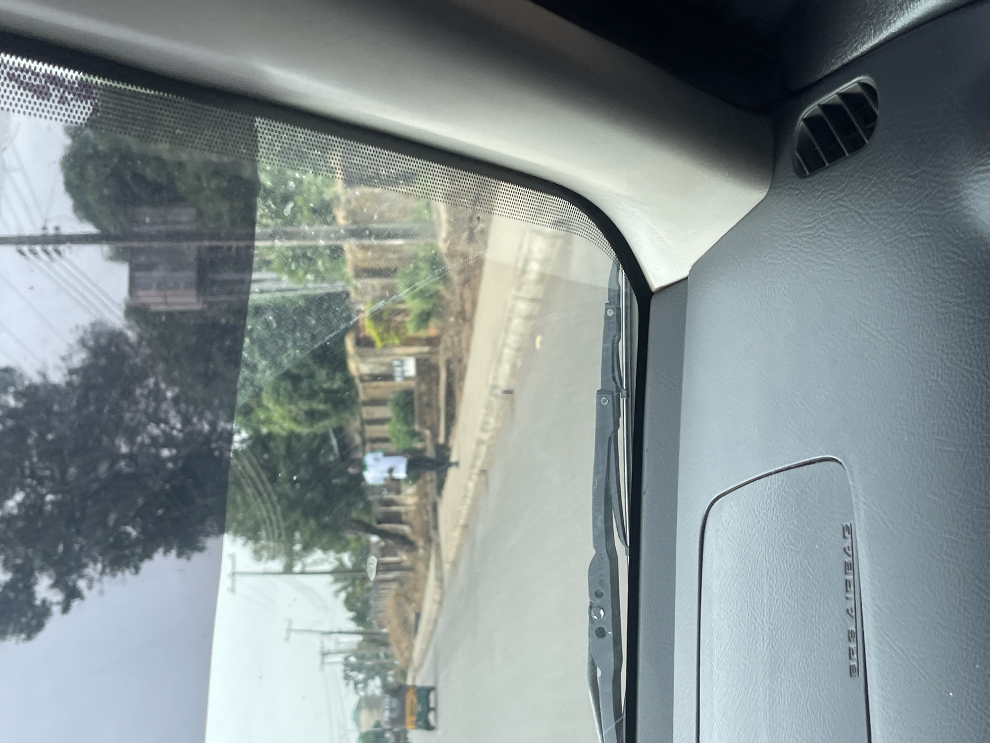 The image is taken from inside a vehicle, looking out through the front windshield. The view through the windshield shows a street scene in Jos, Nigeria. On the side of the road, there are buildings, trees, and telephone poles. The street is paved and appears to be a residential area. In the distance, there is a motorized vehicle (likely a tricycle) traveling on the road. Inside the vehicle, parts of the dashboard are visible, including an air vent and the passenger side airbag cover. The windshield has a wiper attached to it and there is some dirt on the glass.