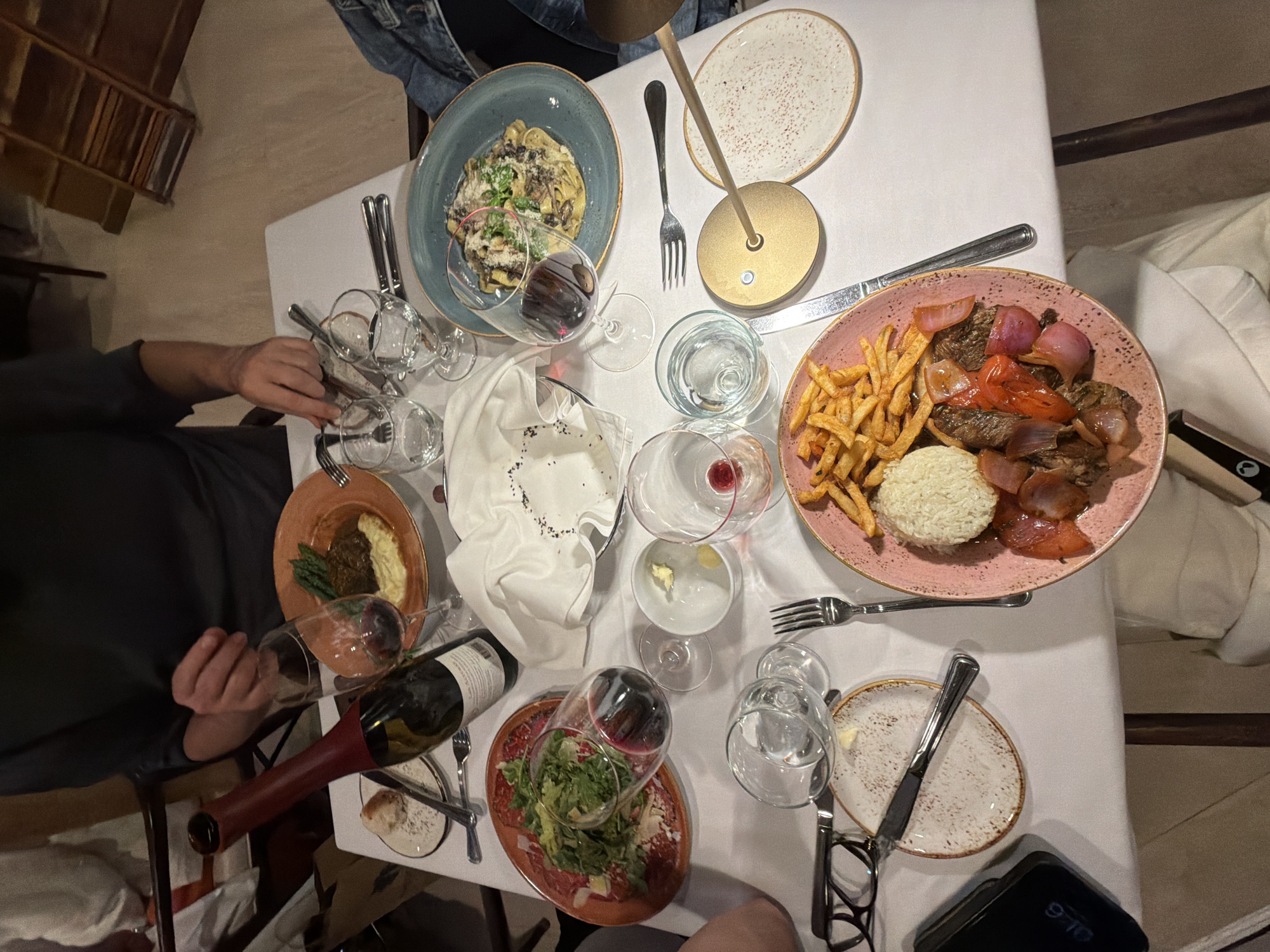 The image captures an overhead view of a dining table, presumably in a restaurant setting. The table is covered with a white tablecloth and is set with multiple plates of food, a variety of glassware, and cutlery. The dishes appear to be different courses or meals. The plates contain salads, meat dishes accompanied by fries and rice, and pasta. There is a tilted bottle of red wine on the table. Several people are seated around the table, their hands and arms visible.
The presence of the restaurant setting and the dining arrangements suggest a social gathering or meal taking place. The timestamp on the phone is visible, 9:10, suggesting the time it was taken. It would be difficult to determine the exact location from visual analysis alone. Based on the tags provided by the user, the location of the event is the city of Key Biscayne, United States of America.