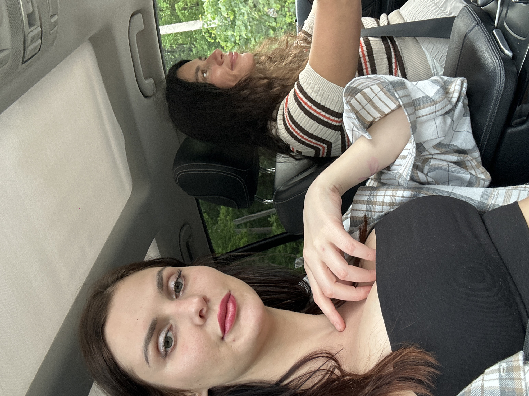Two individuals are seated in a car. The individual in the foreground, on the left, is a woman with long brown hair and makeup. She is wearing a black strapless top and a plaid shirt. She is looking at the camera and has her hand resting on her chest. The individual in the background, on the right, is also a woman with long dark hair. She is wearing a striped shirt and appears to be driving. The car interior is visible, including the dashboard and a sunroof. Outside the car, trees and foliage can be seen. The setting appears to be a suburban or rural area.