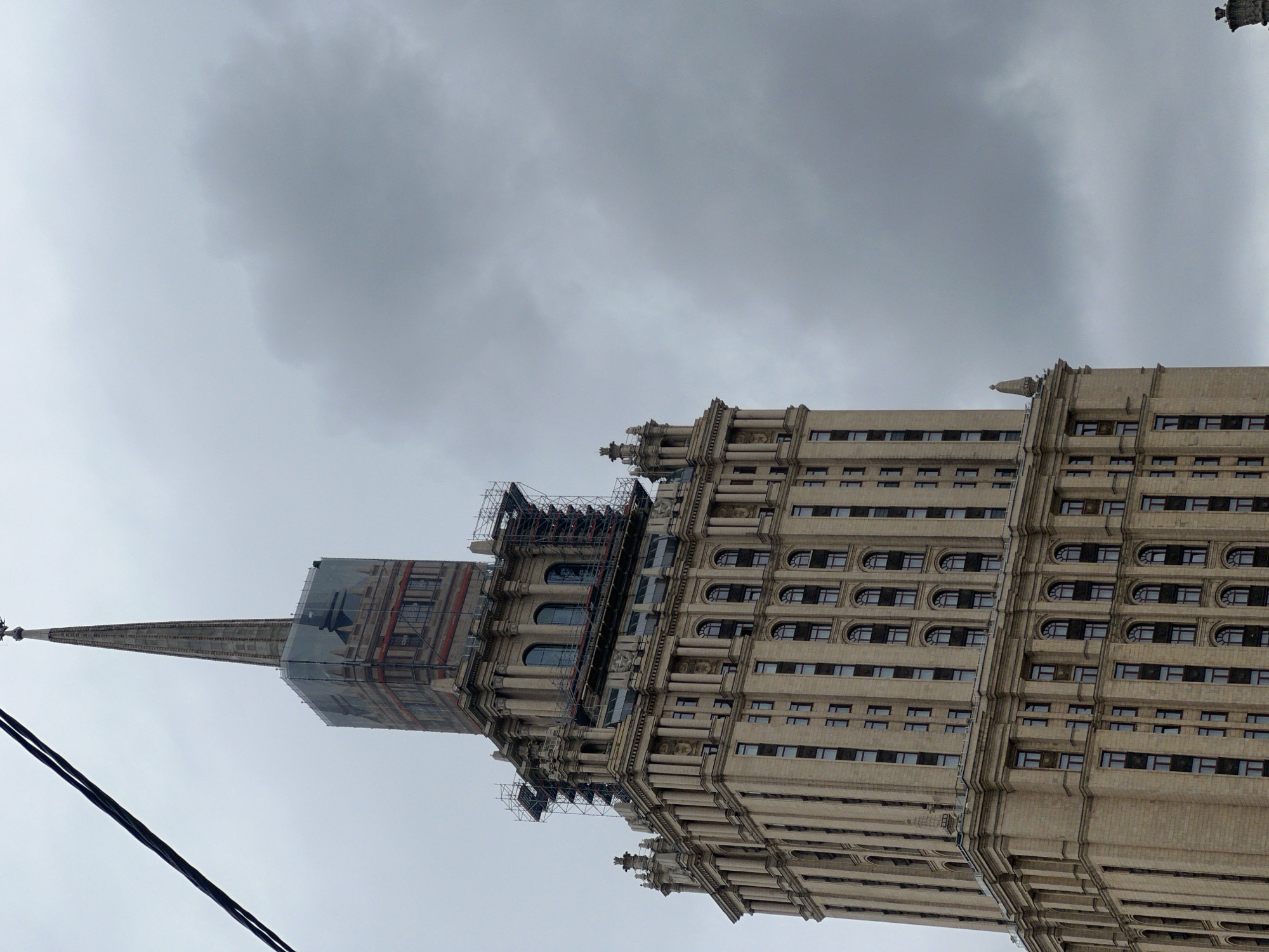 The image captures a section of the Ministry of Foreign Affairs building in Moscow, Russia, under a cloudy sky. The building's exterior is visible, with multiple rows of windows and detailed architectural features. Scaffolding is present on the building. A construction tarp covers the building's spire. Two power lines are visible in the lower left corner of the photo.