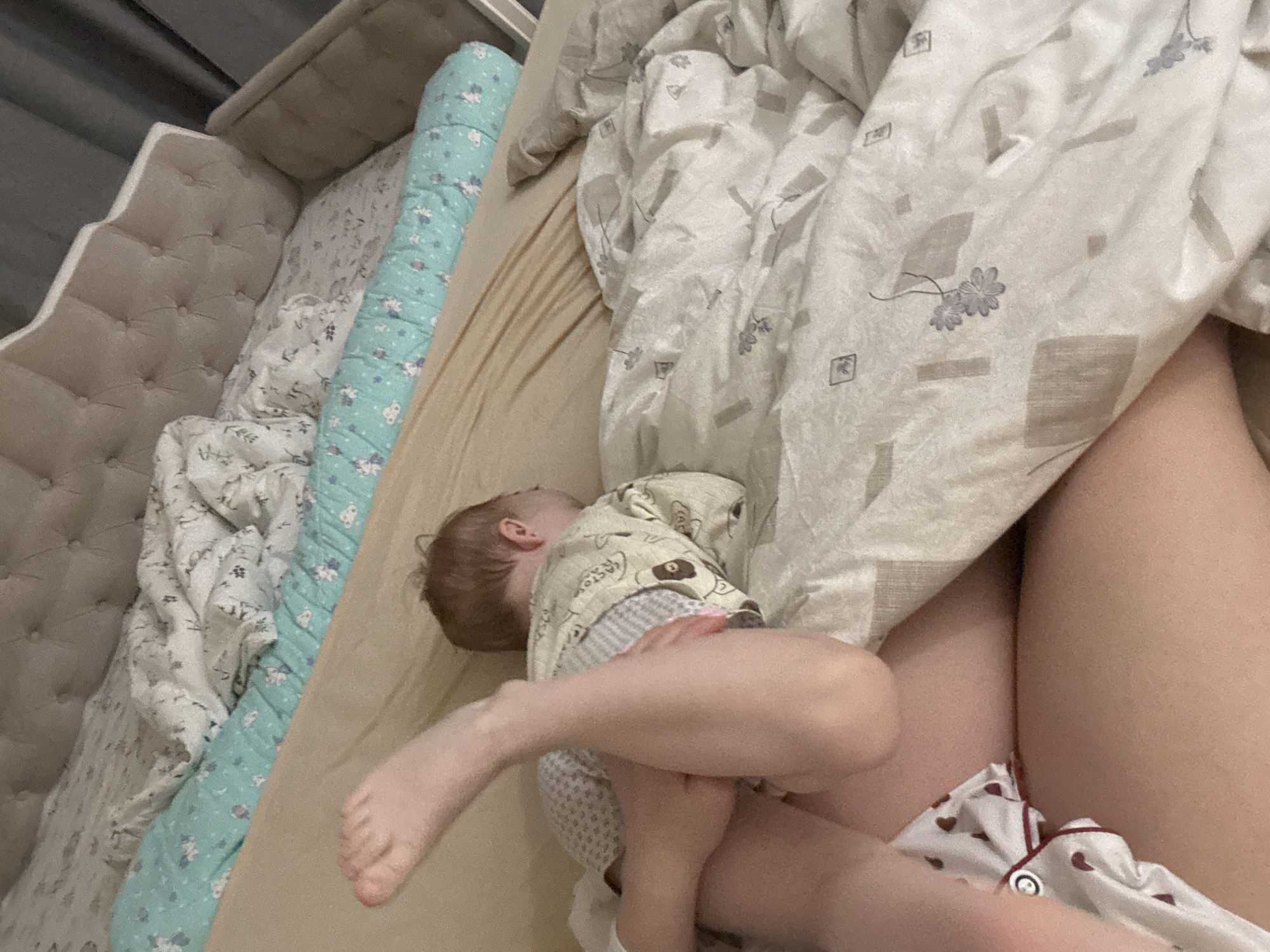 A child lies on a bed next to an adult, with bed linens patterned in neutral tones and a turquoise roll pillow bordering a quilted headboard. The adult wears heart-patterned pajamas, partially obscured by a sheet. The location is a bedroom in Ферма, Russia.