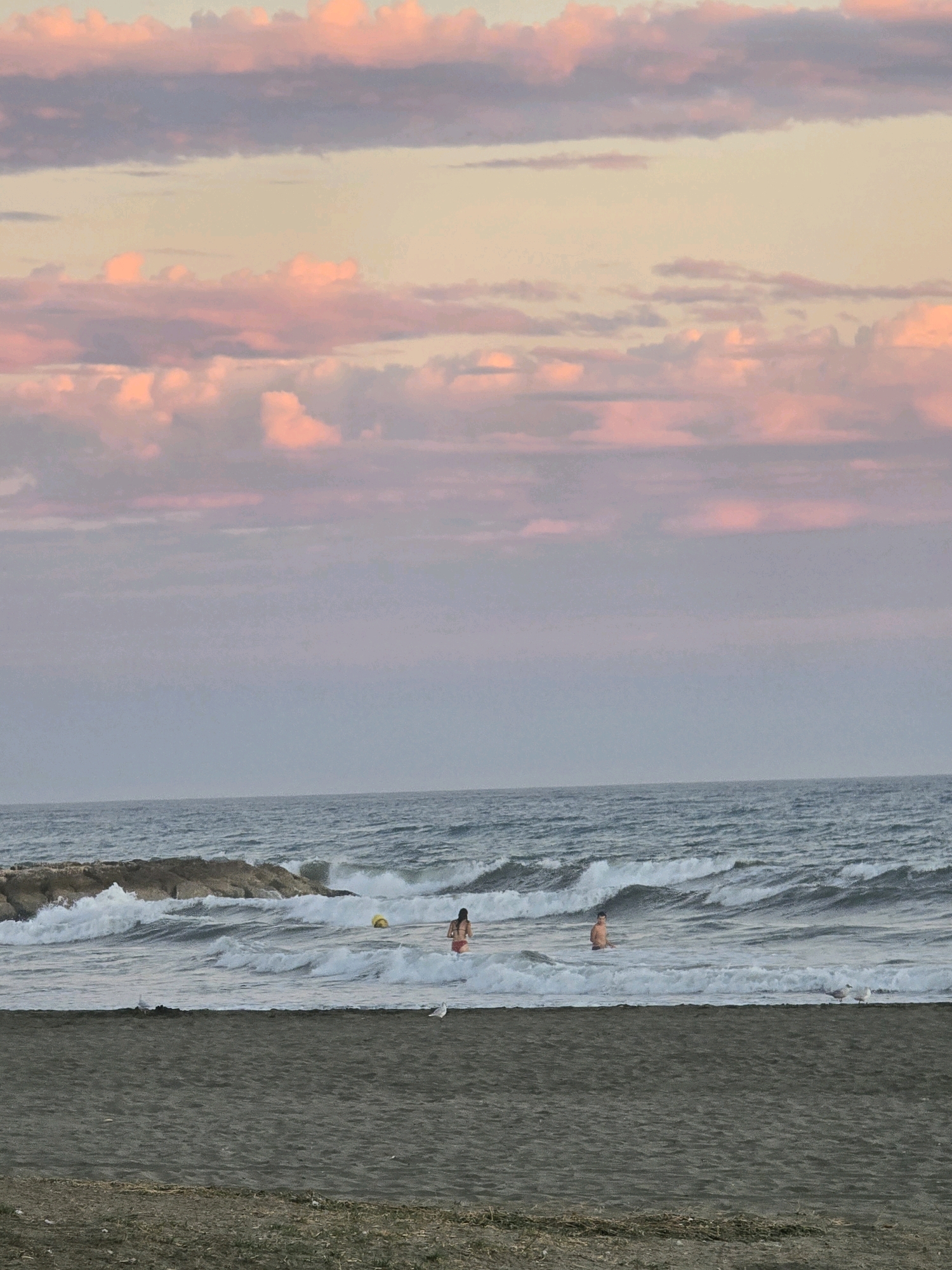 The image captures a serene beach scene at dusk, likely on the coast of Málaga, Spain. A sandy shore occupies the foreground, with sparse vegetation. In the midground, the sea is active with moderate waves crashing against a rocky breakwater. Two individuals, a male and a female, are partially submerged in the water, enjoying the ocean. A yellow buoy is also visible in the sea. Two seagulls are present on the beach. The sky above is painted with soft hues of pink and purple, indicative of a sunset, with scattered clouds adding texture to the twilight atmosphere.