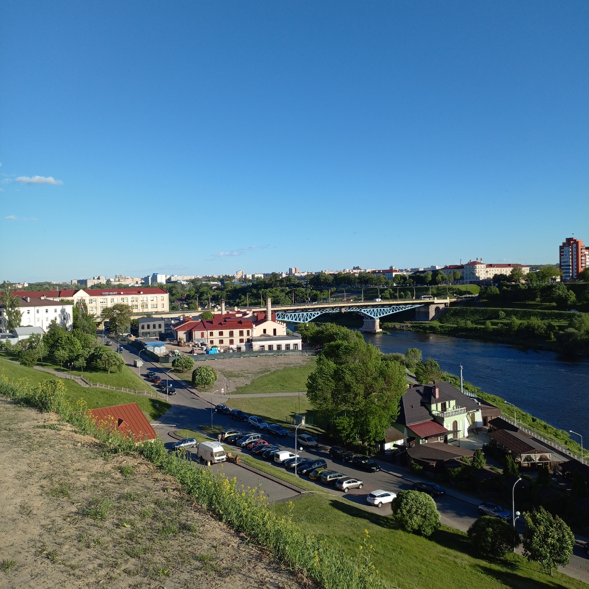 The image depicts a cityscape in Unknown, Unknown, taken from an elevated viewpoint. The sky is a clear blue with a few scattered clouds. In the foreground, there is a grassy slope with a dirt path leading downwards. Patches of yellow wildflowers are scattered along the slope.
Moving into the middle ground, a street runs parallel to the river, with several cars parked along its side. A building with a red roof sits on the edge of the slope. A river flows horizontally across the frame, with a blue bridge spanning it in the middle. Along the riverbank, there are buildings, including a restaurant or cafe with outdoor seating.
In the background, there is a mix of buildings and trees, indicating an urban area. Some buildings are taller than others, with one high-rise apartment building visible on the right side. The architecture appears to be a mix of older and more modern styles. Overall, the scene is typical of a European city with a river running through it.