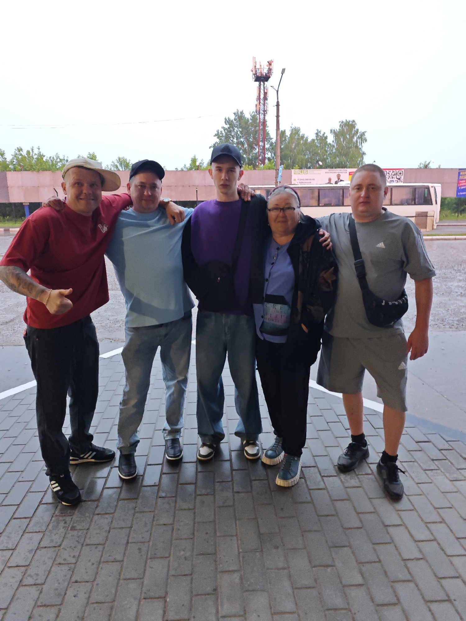 A group of five individuals, three adult males and two adult females, are posing for a photograph in an outdoor setting, likely a bus station, in Chaikovsky, Russia. The ground is paved with rectangular bricks. In the background, there is a white bus with advertisements visible on its side. A tall communication tower and trees are also present in the background. The sky appears overcast.
The individuals are arranged in two rows. In the front row, from left to right, are three adult males. The leftmost male is wearing a red t-shirt, dark pants, a baseball cap, and sneakers. He is giving a thumbs-up gesture. The middle male is wearing a light blue t-shirt, light blue jeans, and dark shoes. He has his arm around the shoulders of the person to his left and the person to his right. The rightmost male in the front row is wearing a grey t-shirt, grey shorts, and black sneakers. He has a black crossbody bag.
In the back row, a woman with glasses is standing between two men. She is wearing a blue shirt and a dark jacket. To her left is a man wearing a dark purple sweatshirt and dark pants. To her right is another man. Both the man in the purple sweatshirt and the man to his right have their arms around the woman. The man to the right of the woman is wearing a grey t-shirt and grey shorts.
The overall mood appears casual and friendly, as indicated by the posing and the thumbs-up gesture.