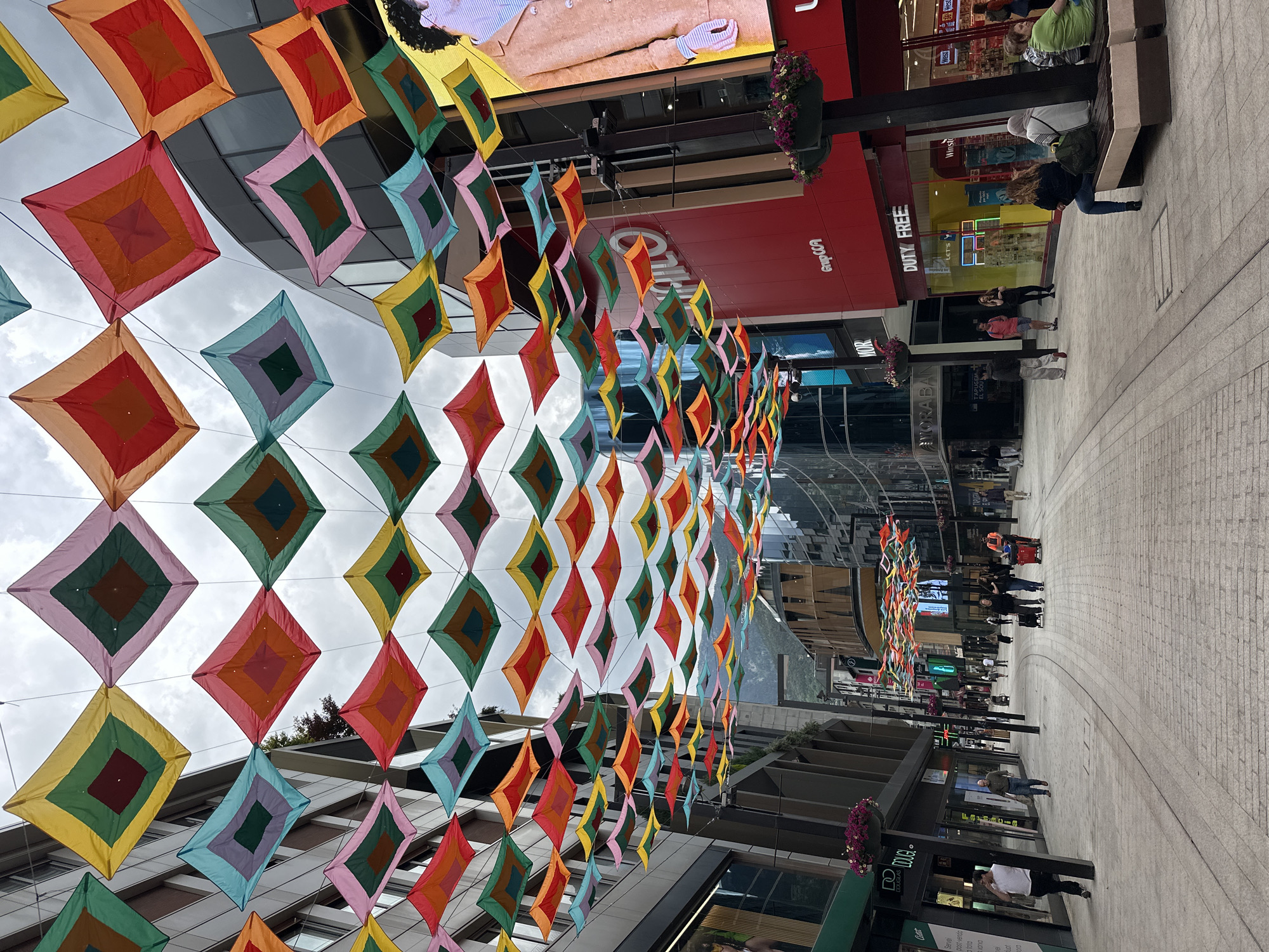 This image captures a vibrant pedestrian shopping street in Andorra la Vella, Andorra, adorned with numerous colorful diamond-shaped fabric decorations suspended overhead. These banners, featuring concentric squares in diverse hues such as red, orange, yellow, green, blue, and pink, create a striking canopy against the overcast daytime sky.
The street is flanked by modern, multi-story buildings housing various retail establishments. On the right side, prominent signage includes "DUTY FREE", "GrupCCA", and "DOUGLAS". Other discernible texts further down this side read "TAPUGEM", "LET'S", "Winsto", and "West". On the left, a sign for "AMORABAN" and "DOUGLAS" is visible, alongside smaller text like "Servei" and "tota". A sign with "EL SOU" is also present.
Several people are seen throughout the scene, engaged in everyday activities such as walking, standing, or sitting on a public bench. The setting suggests a bustling commercial area, characterized by its modern architecture and the festive, artistic street decorations. The weather is overcast, but the bright colors of the decorations maintain a cheerful and lively atmosphere.