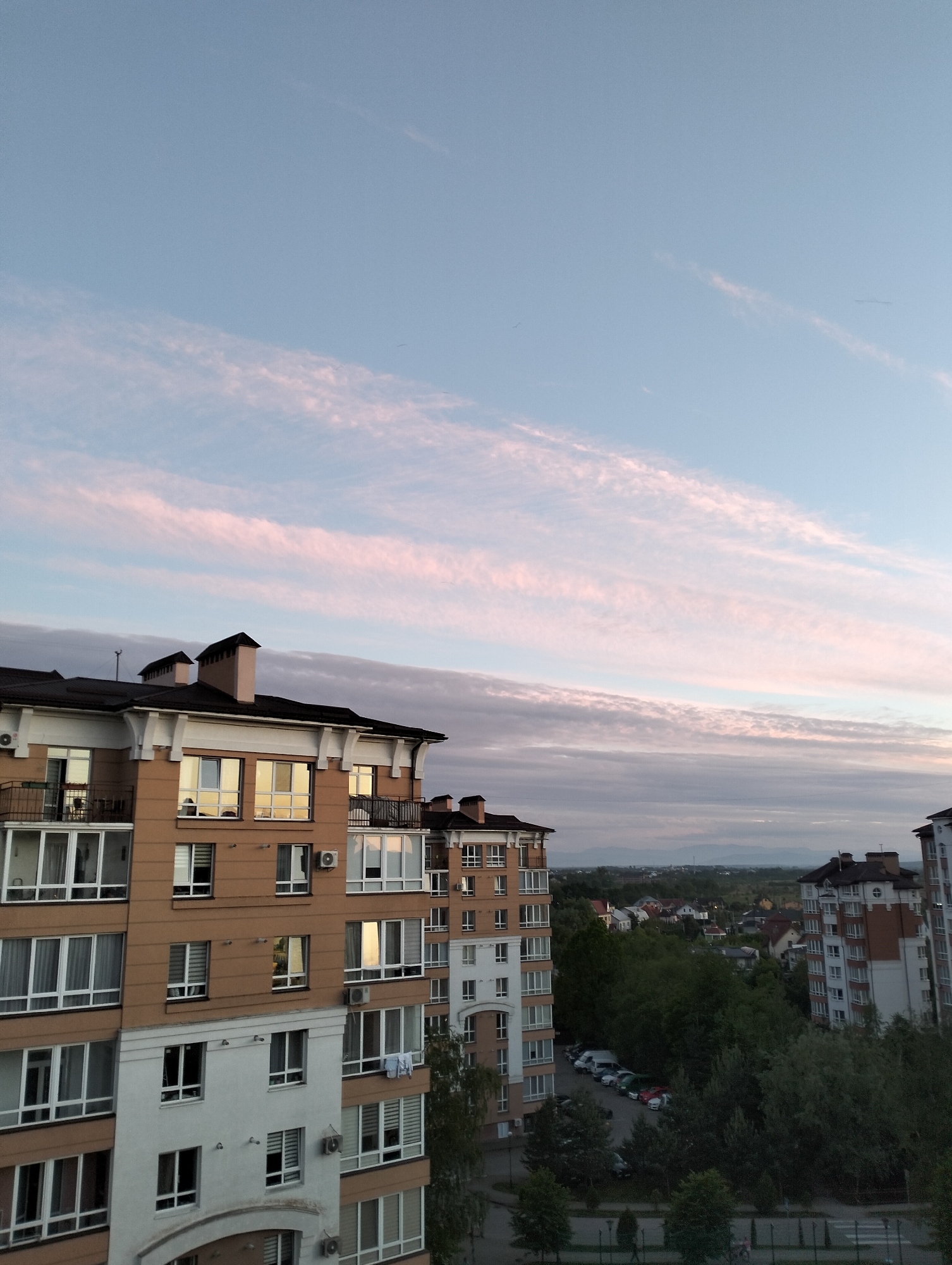 The image captures an urban scene in Krykhivtsi, Ukraine. Two multi-story apartment buildings are prominently featured in the foreground. The building on the left is primarily tan, with white window frames and balconies. The building on the right is brick with white accents and has similar window styles. A few parked cars are visible in a parking area behind the buildings. Beyond the buildings, trees fill much of the middle ground, partially obscuring smaller residential structures. In the distance, a muted mountain range is visible under a twilight sky streaked with pink and grey clouds. The sky transitions from a lighter blue at the top to a greyish hue near the horizon.