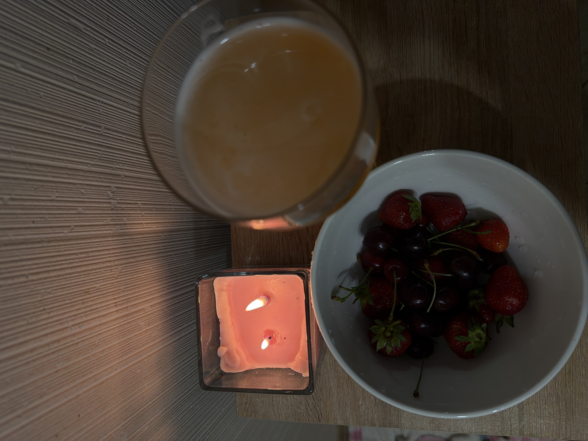 A high-angle shot displays a scene comprising a drink, a bowl of fruit, and a candle. The drink is contained in a clear glass, filled with a beige-colored liquid. Adjacent to it is a white bowl brimming with strawberries and cherries. A lit pink candle in a clear square glass holder sits next to the drink. The objects are arranged on a surface of light-colored wood with visible grain. The location is undetermined and cannot be specified as Cheltuitori, Moldova, based on the visual information.