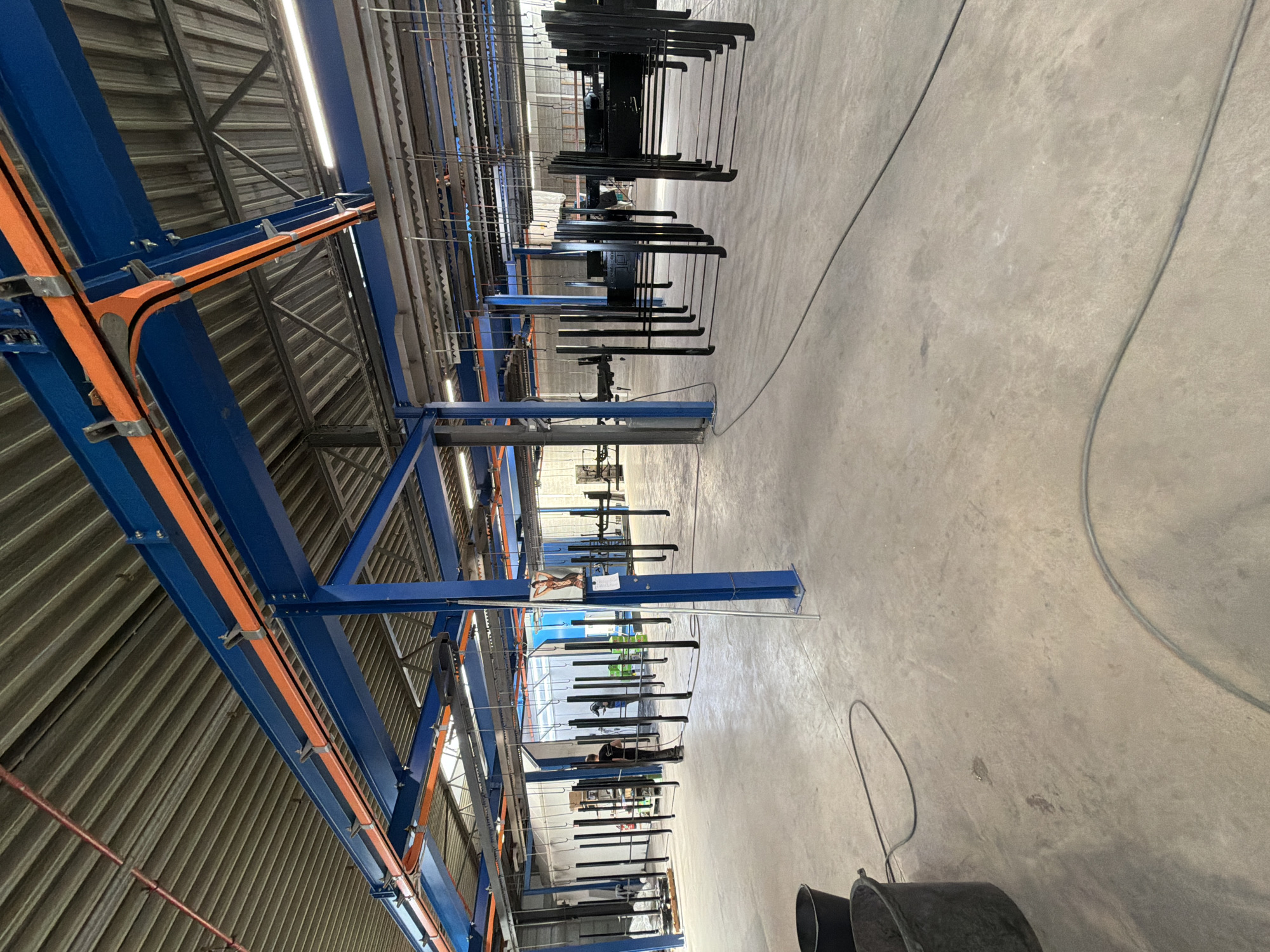 The image shows an indoor industrial setting, likely a factory or workshop, in Mol, Belgium. The scene is dominated by a large blue metal structure, possibly part of an overhead conveyor system or racking. Orange conduits are attached to this structure, running along its beams. The ceiling is corrugated metal, and fluorescent lighting is visible.
Several people are present, appearing to be workers. One individual is visible near the middle of the frame, wearing dark clothing and working on or near the overhead system. Another person, also in dark clothing, is further down and partially obscured by hanging objects.
The primary activity appears to be some form of manufacturing or processing, suggested by the hanging metal parts. These parts, which look like frames or components, are suspended from the overhead system. They are dark in color and appear to be awaiting treatment or assembly. The concrete floor is clean, with some electrical cables running across it.
The time of day is not clearly discernible, but the artificial lighting suggests it's an indoor operation. The weather is irrelevant as the scene is indoors. The overall impression is one of an active industrial environment. No visible text is present in the image.