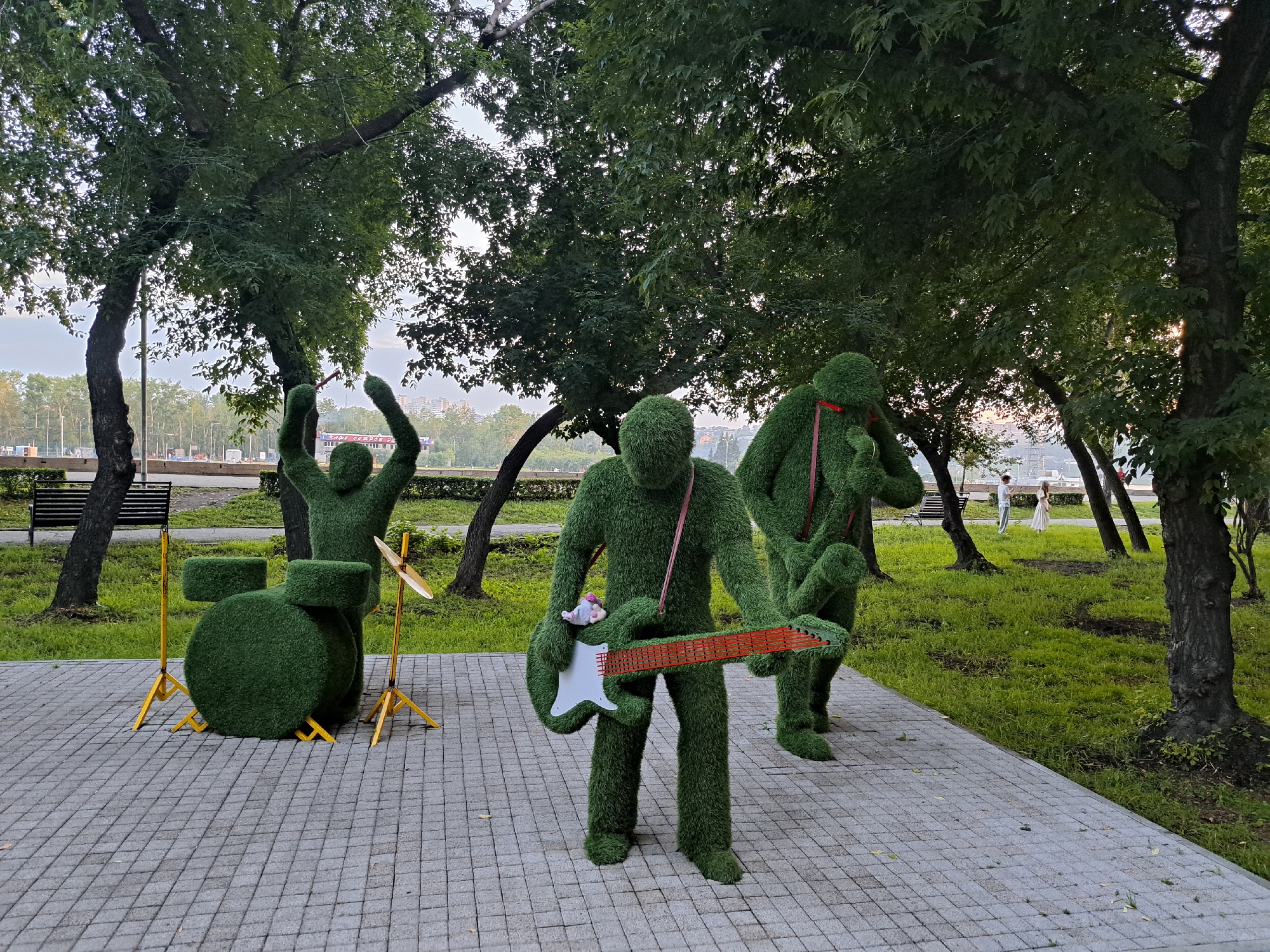 A group of three topiary figures shaped like people are arranged in a park in Irkutsk, Russia. The figures are covered in green artificial grass and are positioned to resemble a band. One figure, in the foreground, holds a white electric guitar with an orange neck. Another figure, to the right, appears to be singing or playing another instrument. A third figure, to the left, is depicted with arms raised, possibly playing drums, as a green drum set with yellow stands is positioned next to it, along with a cymbal. The figures are on a paved walkway. The background shows a grassy area with trees, park benches, and a glimpse of buildings and a road in the distance. Two individuals, one in a white dress and one in a light-colored outfit, are walking in the background. The lighting suggests it is late afternoon or early evening, with some sunlight filtering through the trees.