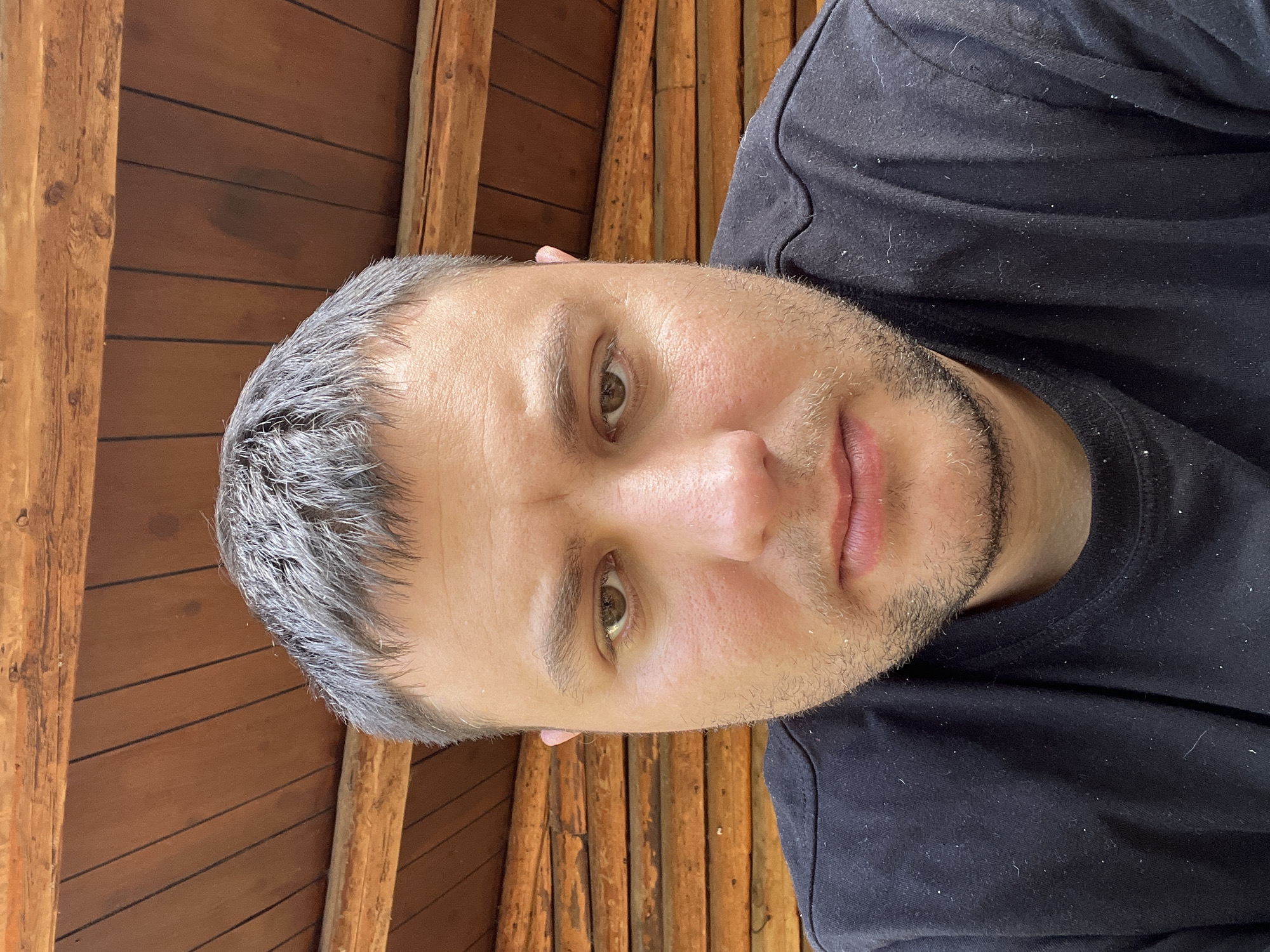 A man with short, graying hair and light stubble is positioned in the center of the frame, looking directly at the camera. He is wearing a black t-shirt. The background consists of a wooden ceiling and walls, constructed from horizontal logs and planks, suggesting an indoor or sheltered outdoor setting. The overall impression is a close-up, candid portrait. The image does not provide any information about the city of Acquaviva, Italy.