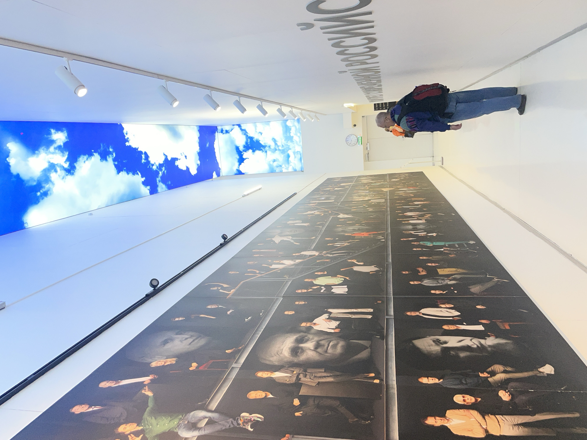 A middle-aged man is seen in an interior location standing on a white tiled floor with walls that are also painted white. The man is wearing blue jeans, a dark jacket over a colored fleece, and a red backpack. He appears to be looking at his phone or a handheld device.
To the left of the man is a large mural comprised of panels, each containing portraits of various people. Above the mural, there are lights on a track. Further to the left is a long rectangular screen showing an image of blue skies and clouds. The text is written on the ceiling above the man and reads “Московской области РОССИИ”. The location is an unidentified exhibition or hall of fame in Khimki, Russia, due to the Russian text. A clock is visible in the background.