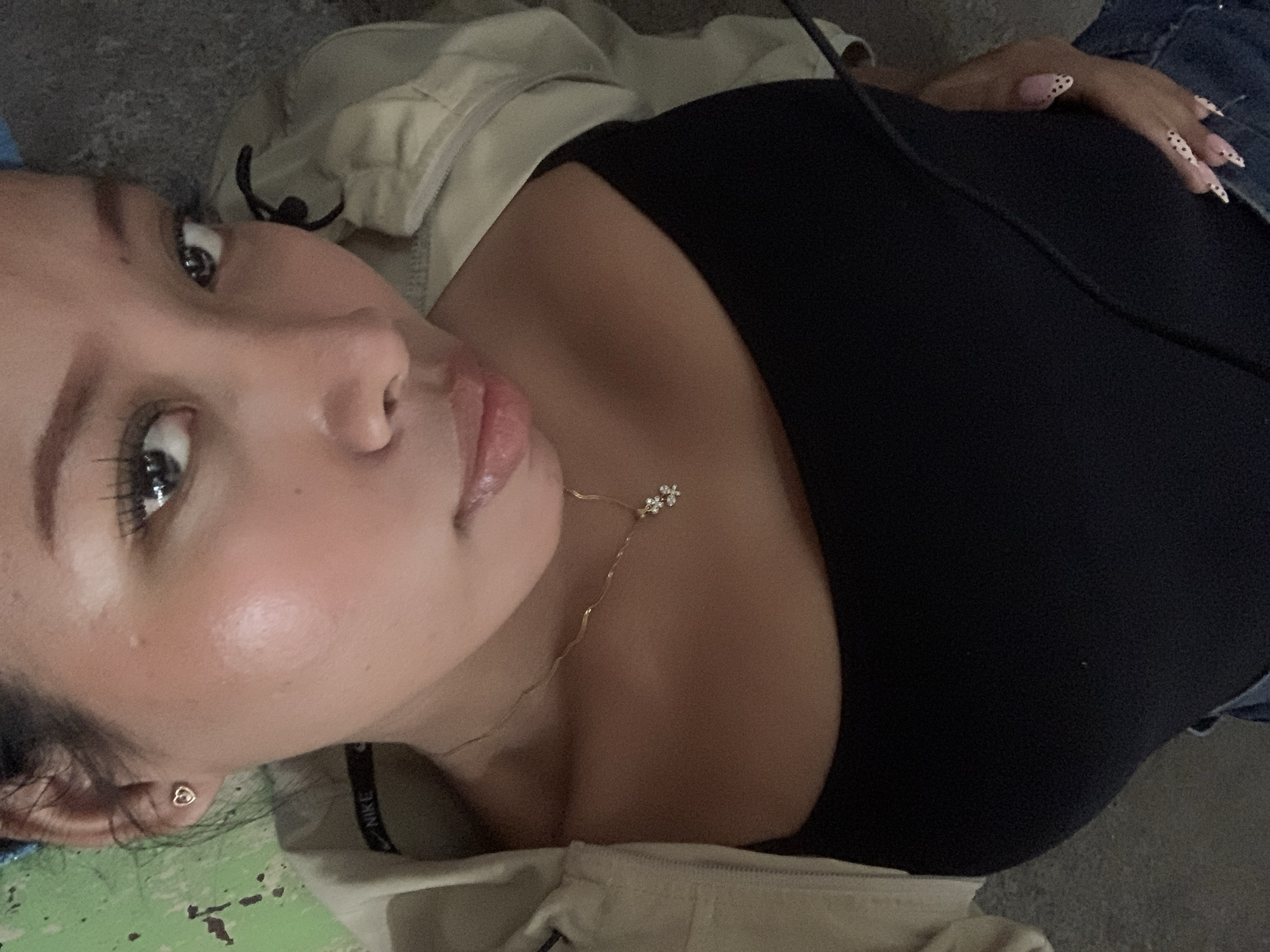 The image is a close-up, high-angle selfie of a person lying down. The person is wearing a black top and a light-colored jacket. A delicate gold necklace with two small flower charms is visible around their neck. Their nails are long and decorated with a pink base and black polka dots. The background appears to be a textured, gray surface, possibly concrete or asphalt, with some green areas visible on the right. The lighting suggests an indoor or shaded outdoor environment. The image does not contain any discernible information that would allow for identification of the location in the city of Tamanbaru, Indonesia.