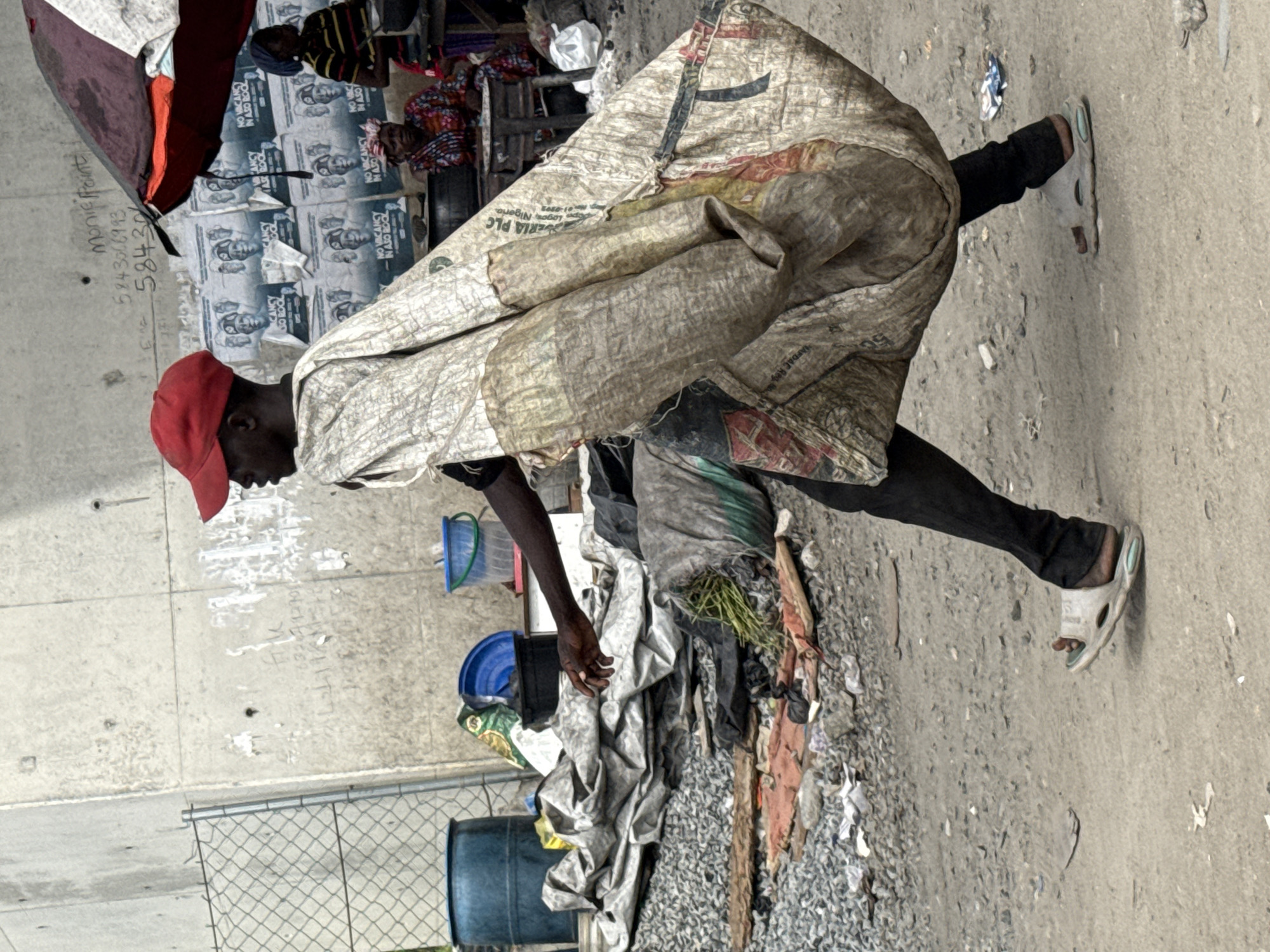 An individual wearing a red cap, dark pants, and sandals stands on a paved surface next to a pile of trash, with an over-the-shoulder covering made from a large sack or bag. The setting appears to be an urban area, as indicated by the concrete wall with graffiti, chain-link fence, and blue barrels. Posters are visible on the wall. OCR text indicates this is Lekki, Nigeria.