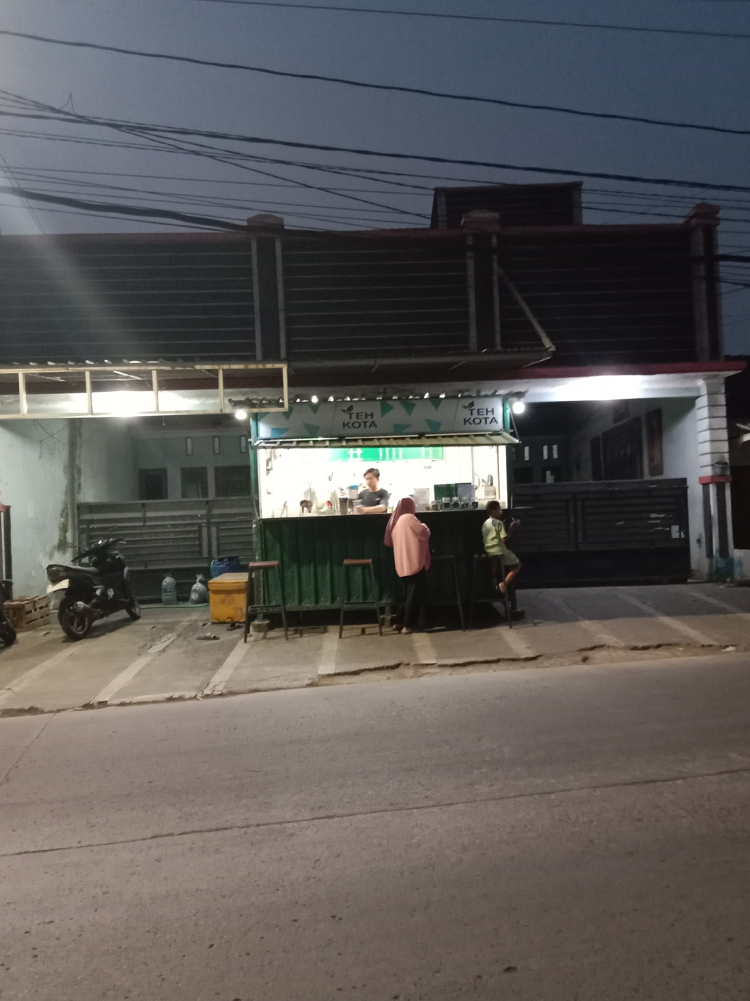 At dusk in Langensari, Indonesia, a roadside food stall known as "Teh Kota" is visible. The stall is constructed from a green shipping container and features a service counter with customers. A male employee stands behind the counter, attending to orders. Two patrons are present: a woman wearing a pink hijab and a young boy in a light green shirt sitting on a stool. A motorcycle is parked to the left of the stall, and various containers and a yellow cooler are scattered on the ground. The scene is illuminated by the stall's interior lights and ambient street lighting. Overhead power lines crisscross the twilight sky, indicating a developed urban or suburban area. The street in front of the stall is paved, and a sidewalk is present.