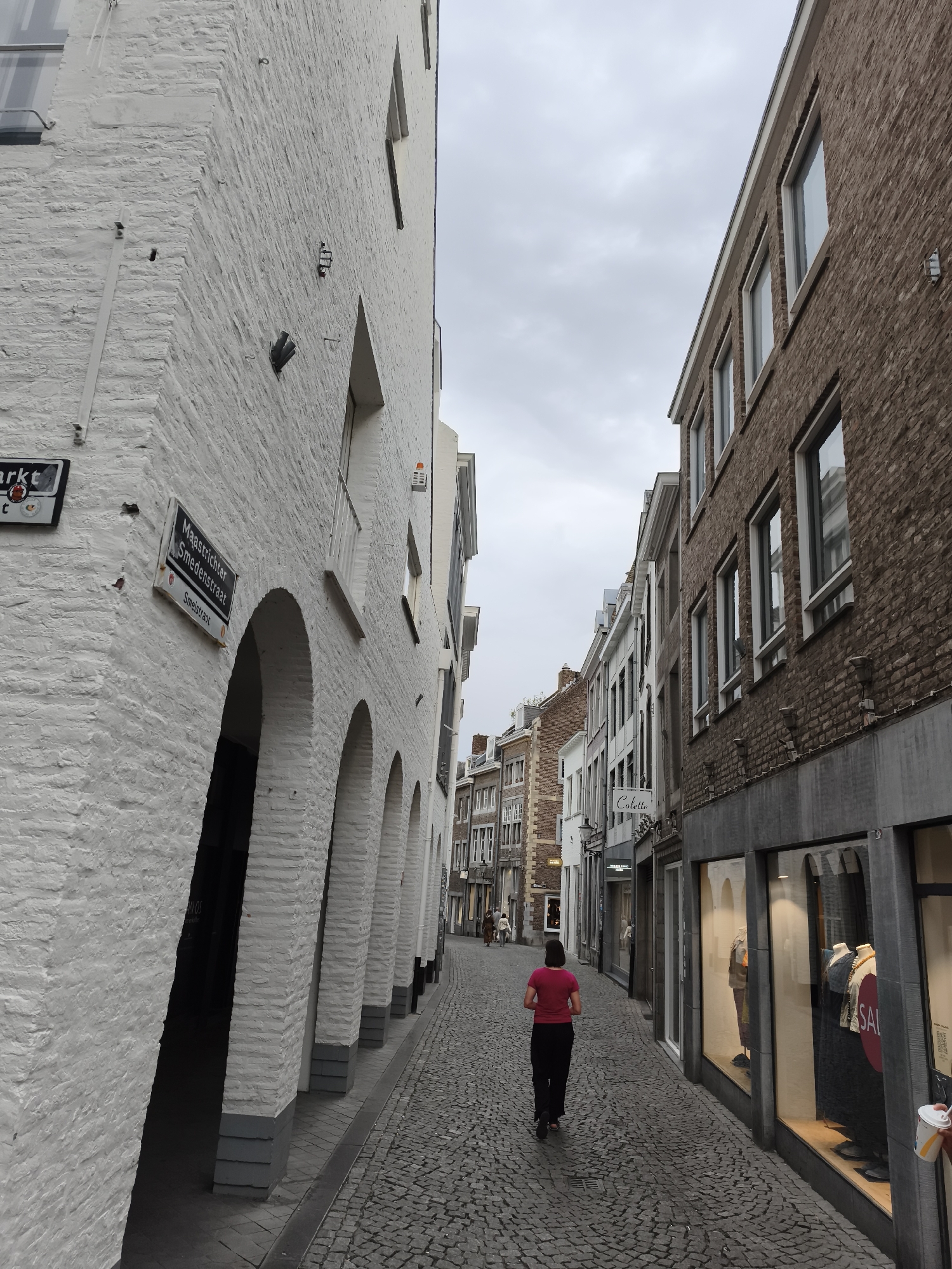The image captures a view looking down a narrow cobblestone street in Maastricht, Netherlands. On the left, a white brick building with arched openings and a sign reading "Maastrichter Smedestraat" is prominent. On the right, a brown brick building with multiple windows lines the street, displaying storefronts. A person wearing a pink shirt and black pants is walking away from the camera down the street. The sky is overcast, and the overall lighting suggests it is daytime. The environment appears to be an urban setting with historical architecture.