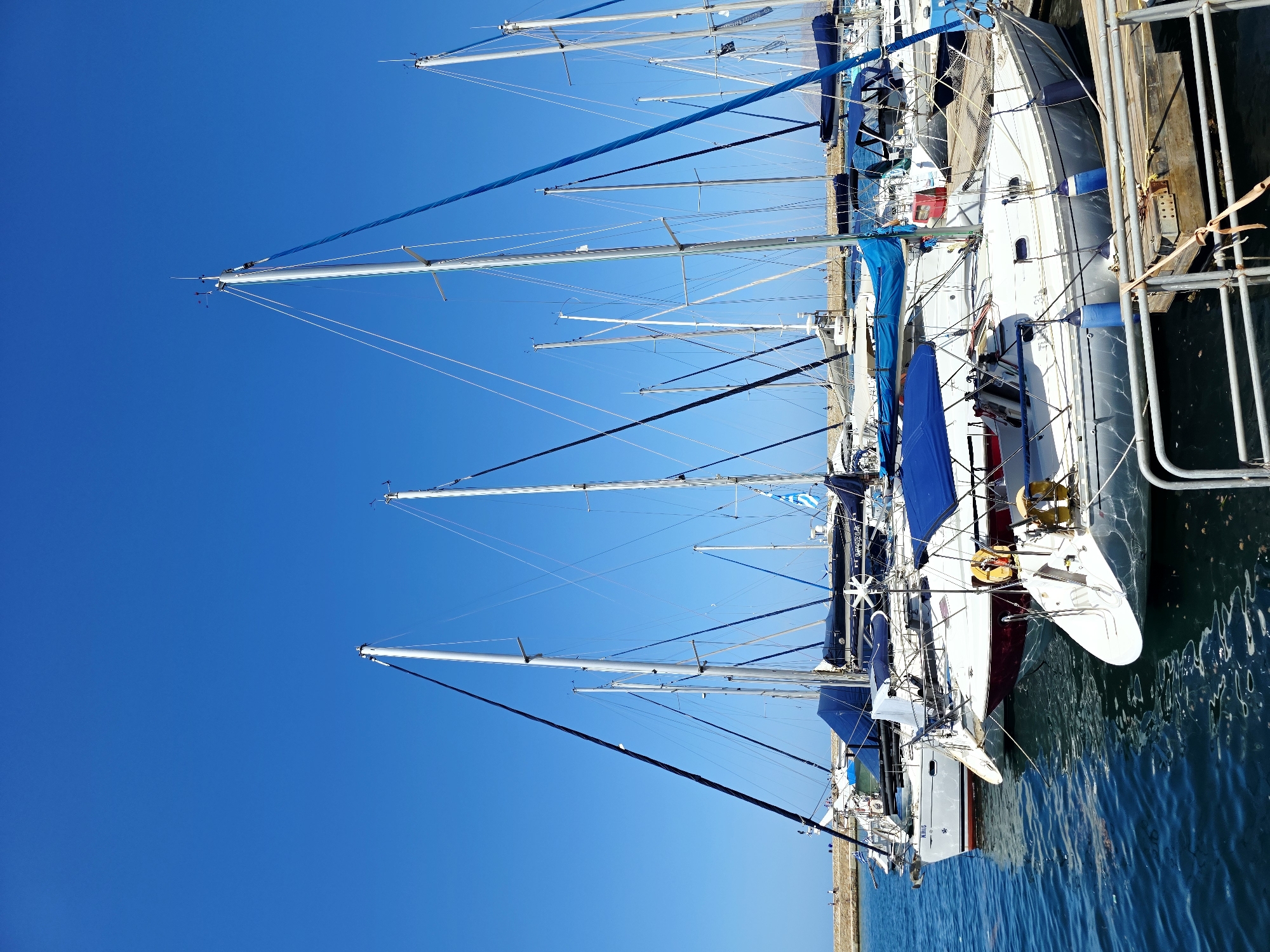 In the city of Chania, Greece, a marina is filled with numerous sailboats. The sky is a clear, vibrant blue, and the water is dark and calm. The sailboats are docked side-by-side, with their masts reaching high into the sky. Some of the boats are white, while others have dark blue covers over their booms. A few people are visible on the boats, tending to them or enjoying the day. In the background, a stone breakwater with a few people walking on it can be seen. The overall scene is one of a peaceful harbor on a sunny day.
