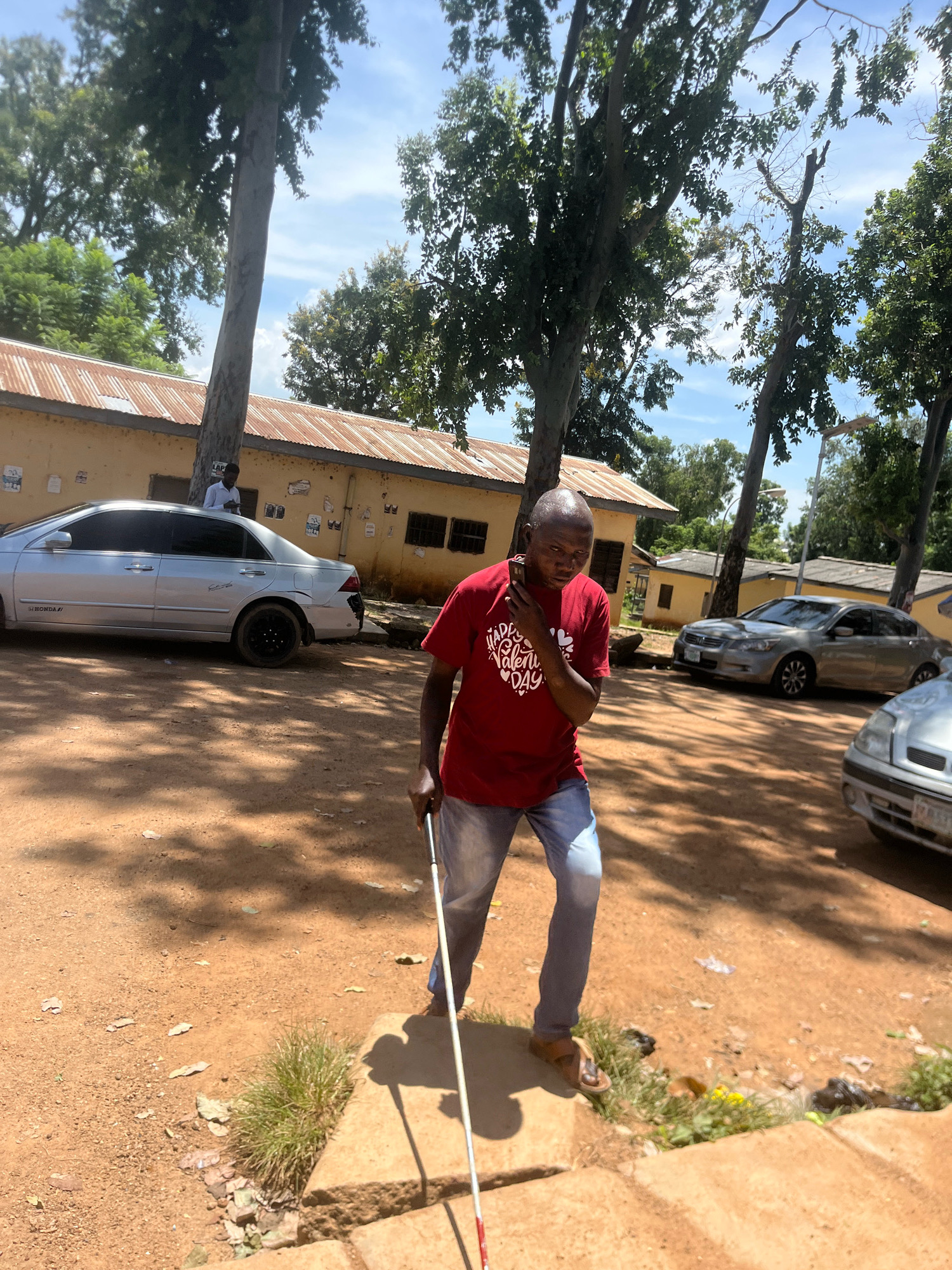The image depicts a man, who appears to be visually impaired, walking with a white cane in what is described as the city of Jos, Nigeria. He is wearing a red t-shirt with a heart graphic and the words "Happy Valentine Day" and blue jeans. The background shows a yellow building with a corrugated iron roof and several trees. Two silver cars are parked in front of the building. The ground is dirt and there are shadows cast by the trees. The overall setting suggests an outdoor environment in a warm climate.
