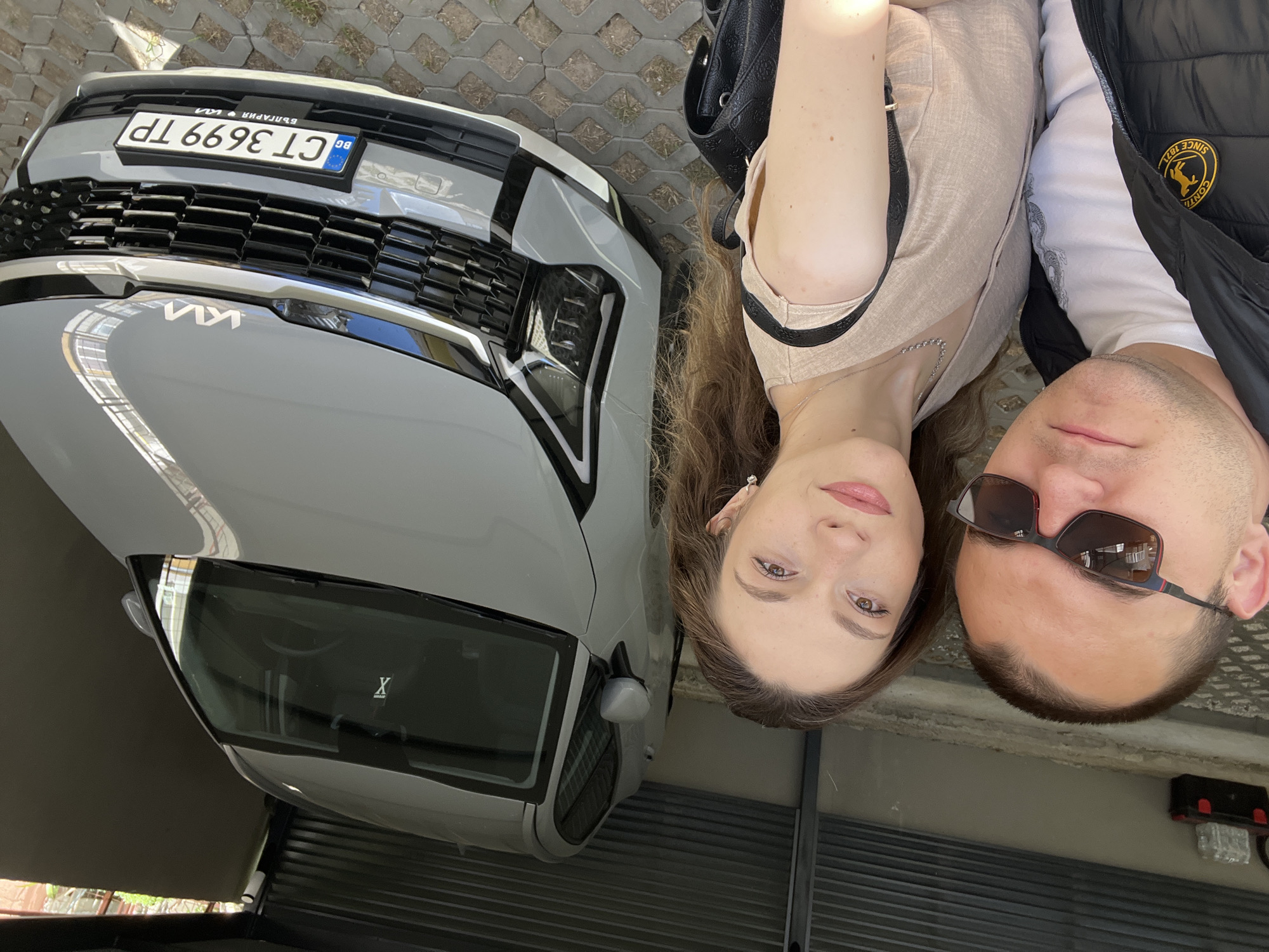 The image depicts a selfie taken by a couple with a gray Kia car. The car's license plate reads "CT 3699 TP" and indicates the car is registered in Bulgaria. The couple is positioned to the right of the car, with the woman on the left and the man on the right, both looking at the camera. The man is wearing sunglasses and a jacket with a patch on the shoulder. The background shows an outdoor paved area and a garage door. The location is not apparent from the image.