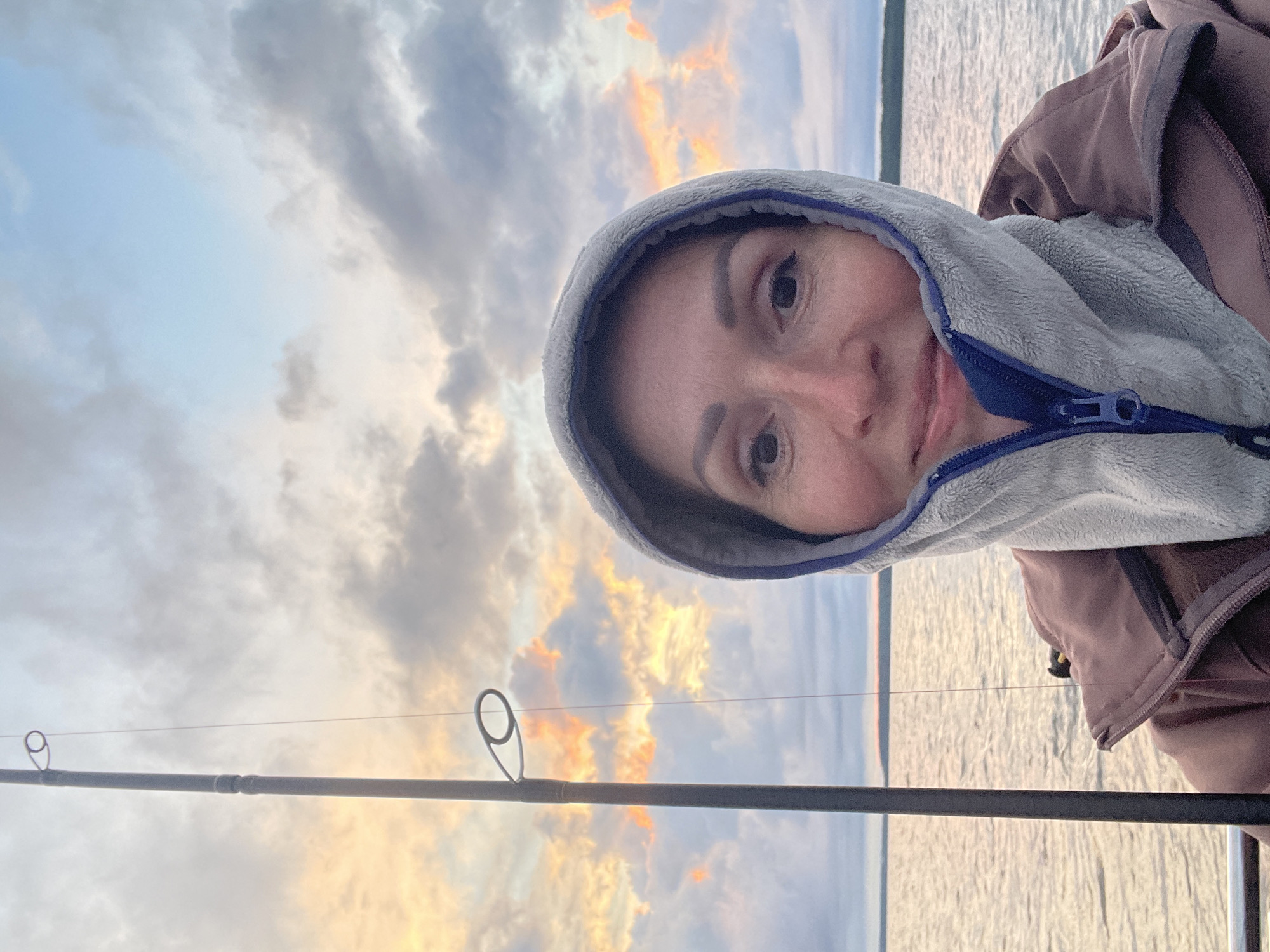 A selfie taken from the perspective of a person wearing a hooded jacket and a fleece hood. The person's face is in the foreground, looking directly at the camera with a slight smile. The background shows a body of water with a faint shoreline and a cloudy sky with hues of orange and pink, suggesting either sunrise or sunset. A fishing rod is visible to the left of the frame, extending upwards against the sky. The image appears to be taken outdoors, possibly on a boat or near a large body of water. There is no discernible location in the city of Yablonovka, Russia, that can be identified from this image.