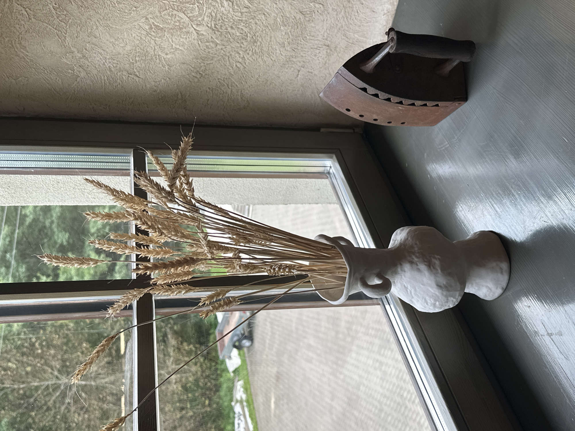 A still life scene is presented on a windowsill, facing outdoors. On the left, a cluster of dried wheat stalks is arranged in a white, irregularly shaped vase with two loop handles. The vase is positioned on a grey windowsill. To the right of the vase, a rusty, vintage iron sits on the same windowsill. The iron is dark brown with a wooden handle and has a distinct triangular shape. In the background, through the window, a paved area is visible, with a small trailer and some greenery beyond. The wall to the right of the window has a textured, beige finish. There are no people or animals present in the scene. The lighting suggests it is daytime. The image appears to be taken from a slightly elevated angle looking down at the windowsill. The specific location in Нарацкі сельскі Савет, Belarus is not determinable from the visual information provided.