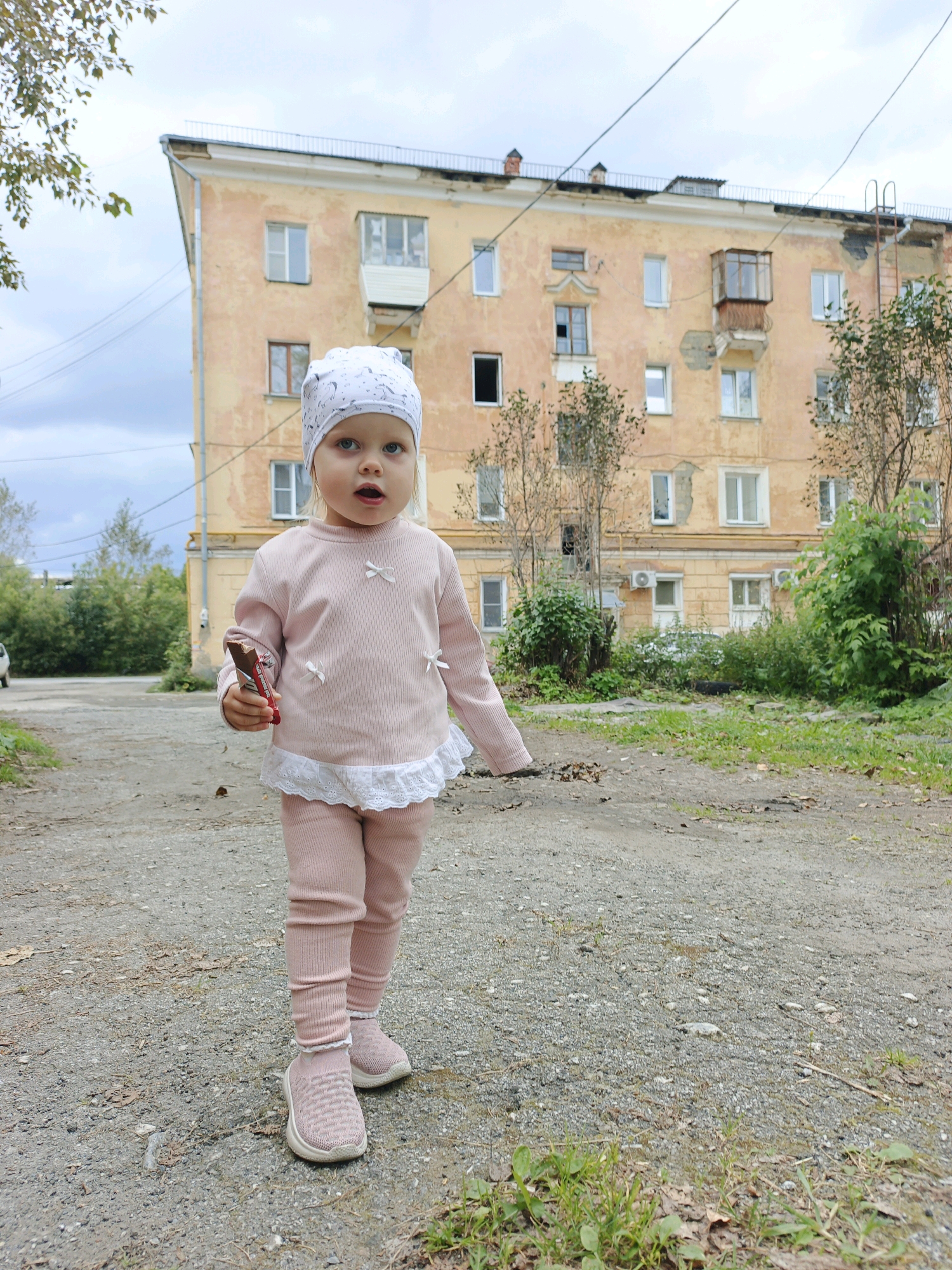 A young Caucasian girl, appearing to be about two years old, stands in the foreground on a gravel path in front of a weathered, orange apartment building. She is wearing a white patterned headscarf, a light pink long-sleeved shirt with decorative bows, and matching pink leggings and sneakers. The girl is holding a piece of chocolate in her right hand and looking towards the camera with her mouth slightly open. The building behind her has multiple windows, some boarded up, and a small, enclosed balcony on the second floor. Overgrown vegetation and trees surround the building. The sky is overcast. The location is Zlatoust, Russia.