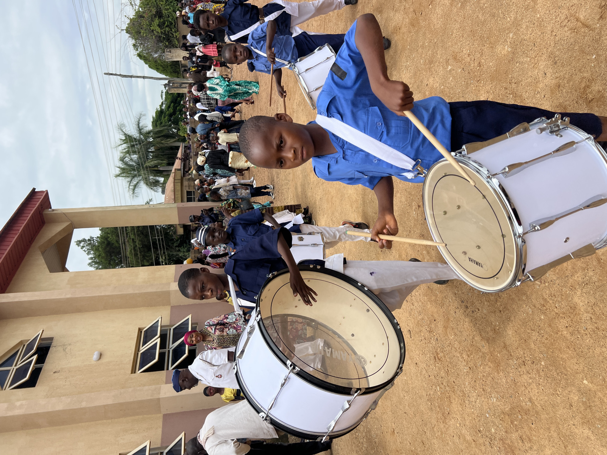 In Jos, Nigeria, on a cloudy day, a group of young boys in blue uniforms march with drums. The boy in the foreground, with a white strap across his blue shirt, holds drumsticks and plays a white bass drum with a Yamaha logo. Another boy behind him, also in a blue uniform and with a white strap, plays a similar drum. A larger bass drum, also white, is carried by a boy to the left, who is partially obscured by it. The boys are on a dirt path, with a beige building with open windows in the background. A crowd of people, some in colorful attire, watches the procession. The atmosphere appears festive, possibly a parade or ceremony.