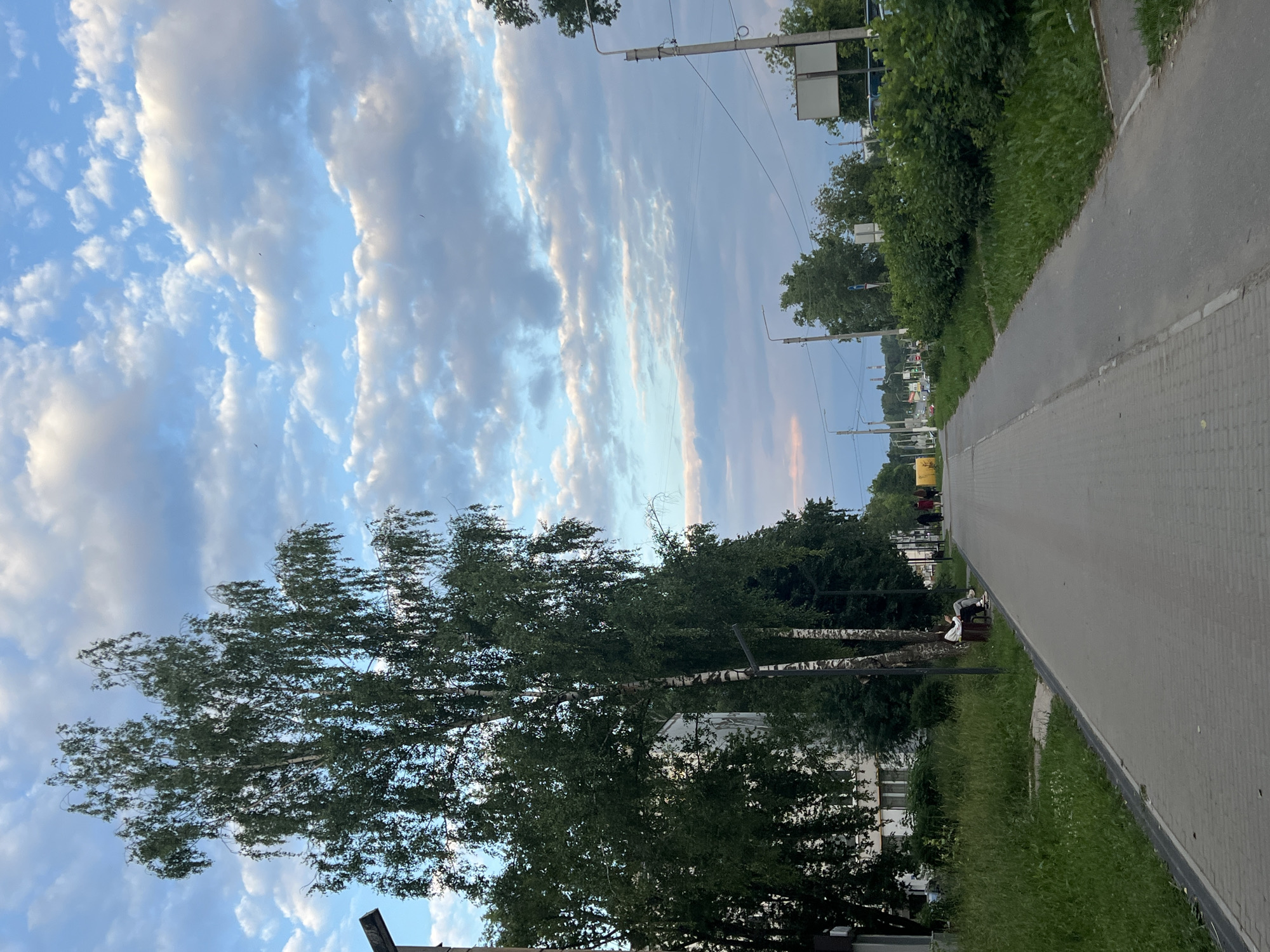 The image captures a wide view of a pedestrian walkway in Chaikovsky, Russia, bordering a park-like area and a residential building on the left. The walkway is paved with gray rectangular blocks and has a distinct gray curb. On the left side, tall green grass grows abundantly along the edge of the walkway and the base of a multi-story white apartment building. A large tree with green foliage and a white trunk stands prominently on the left side of the walkway.
Further down the walkway, a park bench is visible with two individuals seated on it. They appear to be engaged in conversation or simply resting. Beyond them, the walkway continues, leading towards a street with several more people walking. The environment features several utility poles with electrical wires, streetlights, and some signage. On the right side of the walkway, a row of lush green bushes lines the path, and beyond them, more trees and some structures are visible, including what appears to be a bus stop with a shelter.
The sky above is a mix of blue with scattered white and gray clouds, suggesting it is either early morning or late afternoon. The lighting indicates a slightly diffused sunlight, casting soft shadows. The overall scene portrays a quiet, everyday urban environment with elements of nature integrated into the cityscape.