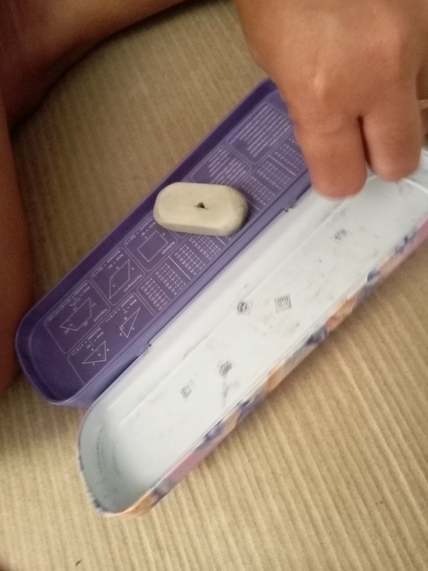 A person's hand is seen hovering over an open pencil case. The pencil case is purple and white, with diagrams and formulas for geometric shapes printed on the purple lid. A light-colored eraser is visible inside the pencil case, resting on the purple lid. The background consists of a textured surface, possibly a mat or carpet. This image was taken in Batam, Indonesia.