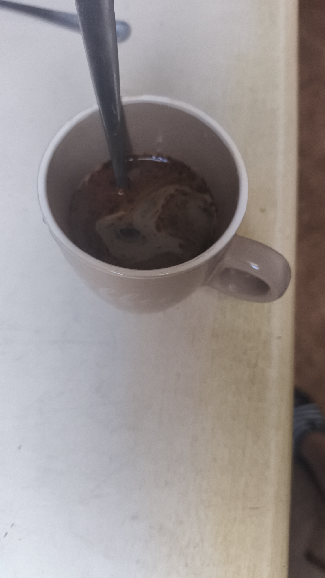 A top-down shot shows a beige mug filled with coffee and a spoon. The mug sits on a beige countertop. Part of a person's foot, wearing a striped sandal, is visible in the bottom right corner. The scene is indoors. There's no indication of the location being in Nowy Tomyśl, Poland.