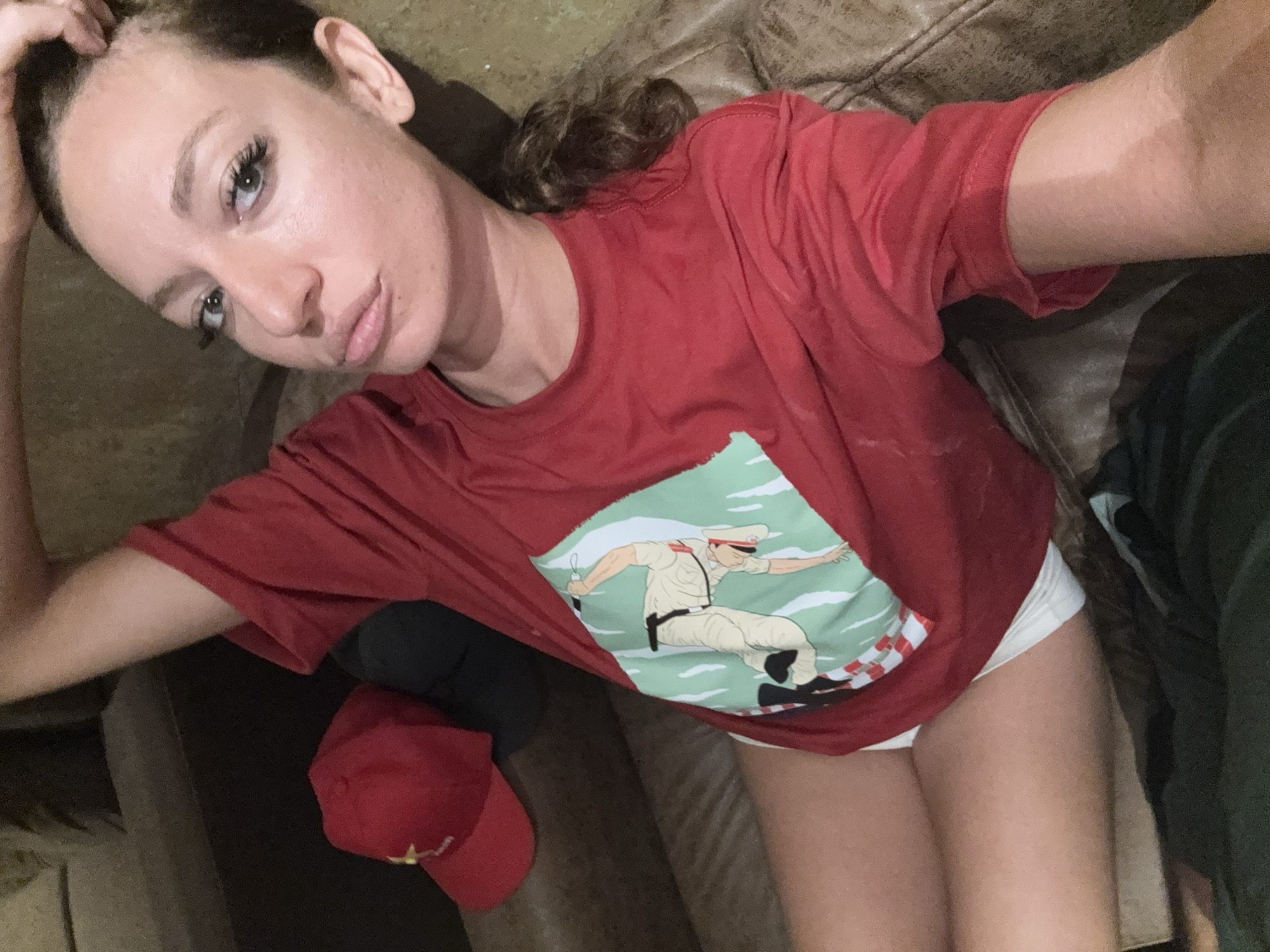 A fair-skinned woman is lying on a brown leather couch, taking a selfie. She is wearing a red t-shirt with a graphic of a man in a white uniform on it and white underwear. A red baseball cap sits next to her. The location is unknown, but the user has stated that it is in Hà Nội, Vietnam.