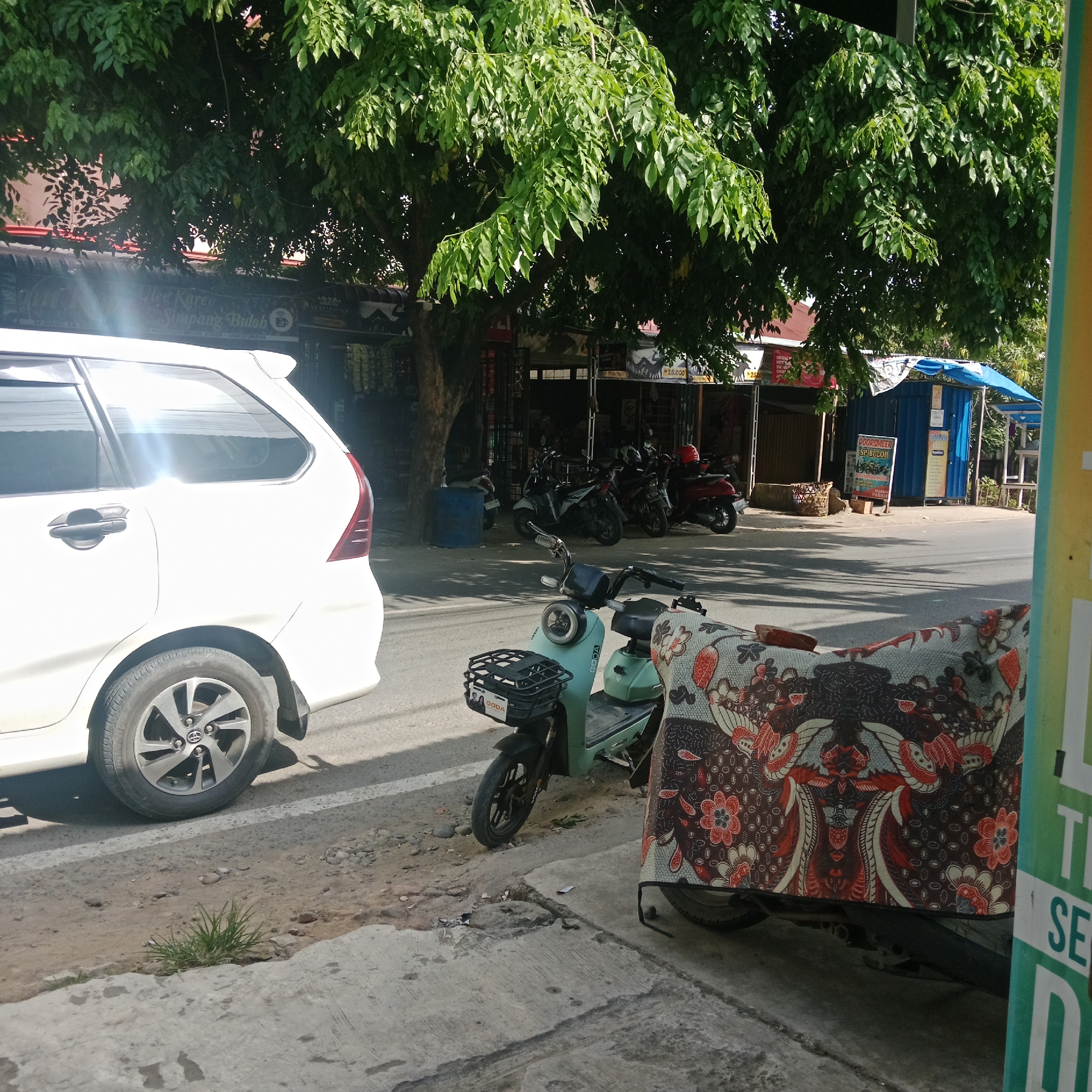 This image captures a bright, sunny daytime street scene in Lhokseumawe, Indonesia, characterized by local commerce and parked vehicles. The setting appears to be a bustling commercial area, possibly along a main road, with various shops or stalls lining one side.
In the foreground on the left, the rear-left portion of a white multi-purpose vehicle (MPV), identified by a Toyota logo on its wheel, is parked beside the road. Its surface reflects bright sunlight. To its right, a light blue electric scooter, branded "GODA" on its front basket, is parked on the paved sidewalk. In the immediate foreground on the right, another scooter is partially visible, largely covered by a vibrant, intricately patterned fabric reminiscent of Indonesian Batik designs.
Behind these foreground vehicles, a row of motorcycles and scooters are parked along the street in front of various establishments. These shops are shaded by a large, leafy green tree that dominates the upper part of the image, casting shadows across the road and sidewalk. Visible elements of the shops include awnings, a blue barrel near the tree trunk, and a blue metal container-like structure with a blue tarp awning further down the street.
Signs provide strong location cues. Partially obscured by the tree, a sign mentions "Kopi Simpang Buloh" (Simpang Buloh is a locale in Lhokseumawe). Another prominent white and blue sign clearly reads "DOORSMEER SP. BULOH" and "BEBAS PARKIR" (Free Parking). Several other signs display "Rp 25.000," indicating prices in Indonesian Rupiah. The general atmosphere suggests a typical daily scene in a Southeast Asian town. No people are explicitly visible in the frame, but the parked vehicles and open shops imply active human presence and commerce.