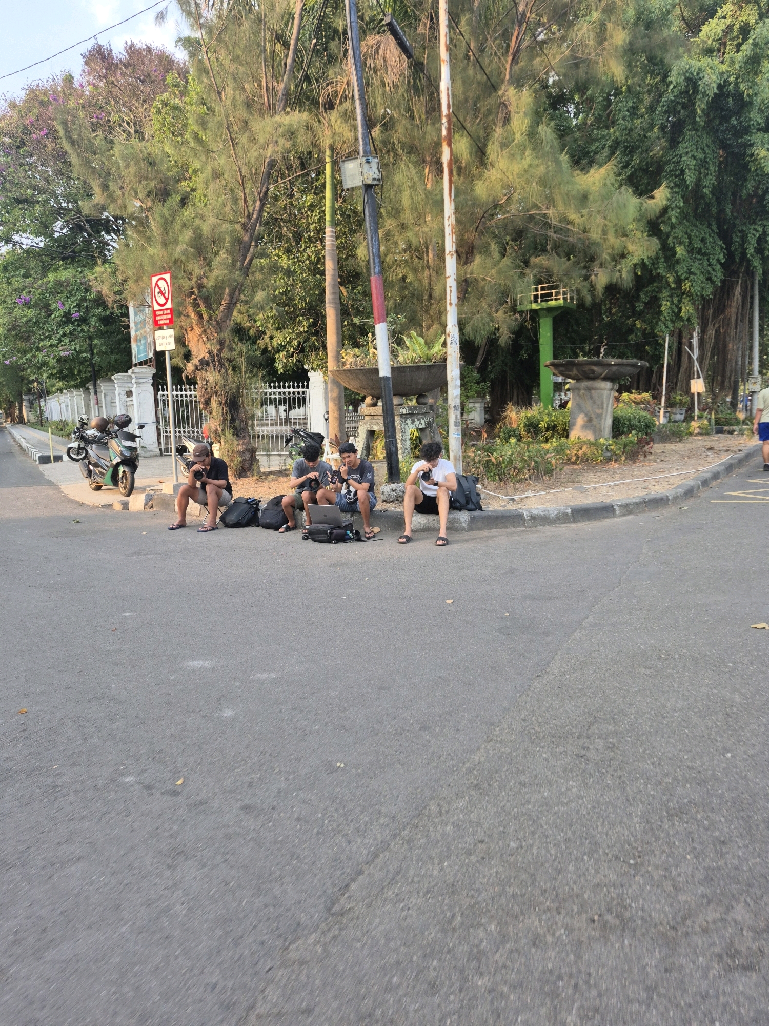 The image shows a street scene in Surakarta, Indonesia, with four young men sitting on the curb. The men are casually dressed and appear to be engaged in conversations or resting. One man on the left is holding a camera and looking at it. The second man from the left is also holding a camera. The third man from the left has a laptop on his lap and is looking at it. The fourth man from the left is holding a camera. There are motorcycles parked on the side of the road behind the men, and a fence with white pillars in the background. The environment includes trees, a streetlamp, and a decorative fountain. The road surface is asphalt, and there are fallen leaves on the ground. The lighting suggests it is daytime, possibly late afternoon given the warm tones.