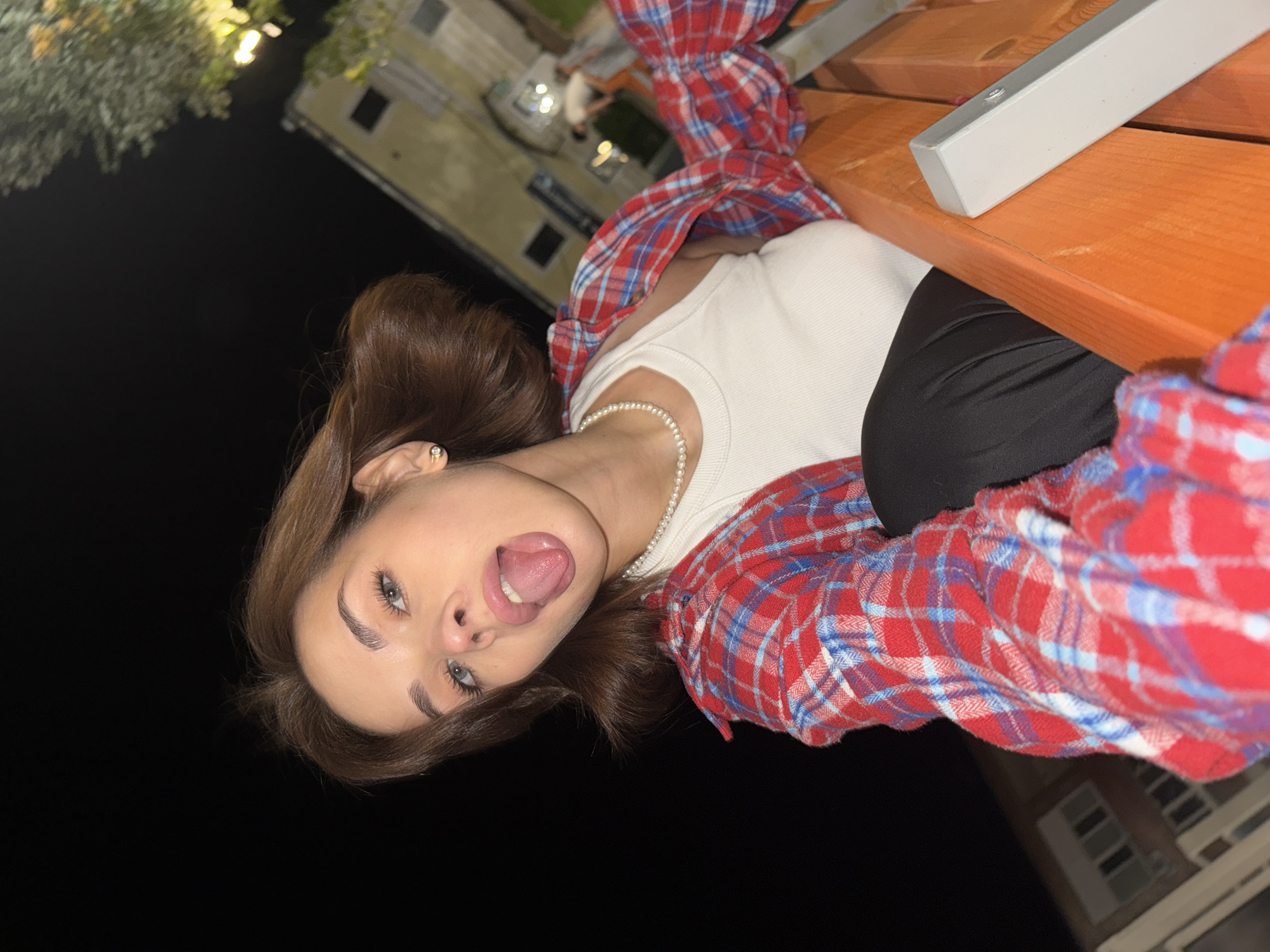 A young woman with brown hair and fair skin is posing for a selfie outdoors at night. She is wearing a white tank top, a pearl necklace, and a red and blue plaid shirt. She is sticking her tongue out and looking at the camera. She is sitting at a wooden picnic table. The background is dark, but there are some buildings and trees visible in the distance. The image was taken in Sarapul, Russia.