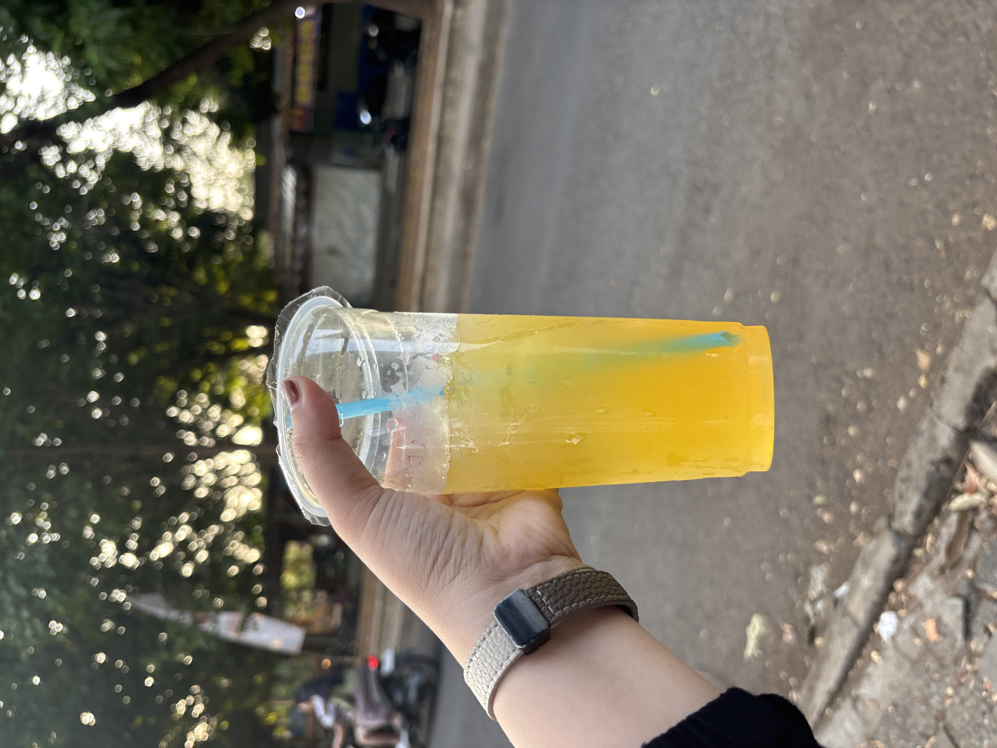 A person's hand, wearing a watch with a beige and grey band, holds a clear plastic cup filled with a yellow liquid and a blue straw. The cup is positioned in the foreground, with the hand extending from the left side of the frame. The background is blurred, showing a street scene with trees and a road. In the distance, a person is riding a motorcycle. The overall impression is of a casual moment outdoors, possibly in a city setting. The presence of a motorcycle and the style of buildings suggest an urban environment in Indonesia, consistent with Cirebon.