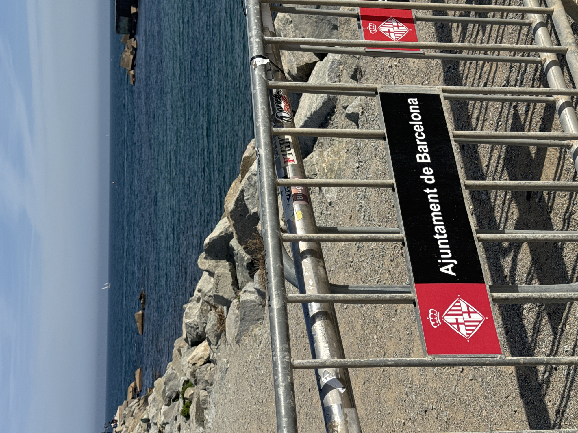 The image captures a scene at the waterfront in Barcelona, Spain. A metal stairway and railing are prominently featured, with an "Ajuntament de Barcelona" (Barcelona City Council) sign attached. The sign has the Barcelona coat of arms on a red field. The stairway leads down to a rocky shoreline where waves lap against the stone. In the distance is a calm sea, with a hazy sky above.