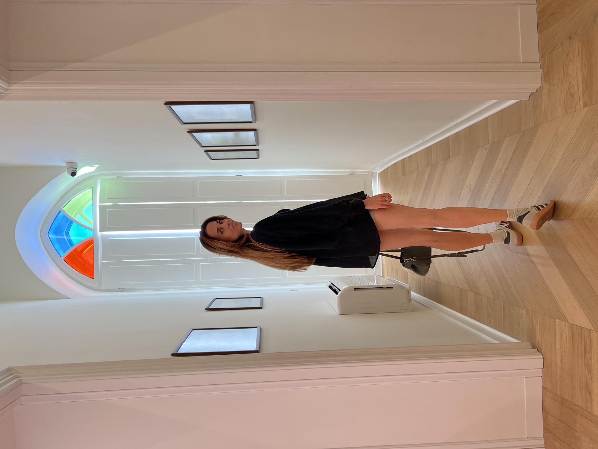 A woman with long, brown hair is standing in a brightly lit hallway with white walls and a light wood herringbone floor. She is wearing a black oversized shirt, denim shorts, white socks with black stripes, and athletic shoes. She is holding a dark green handbag and looking over her left shoulder at the camera. The hallway features framed artwork on the walls and a white air conditioning unit mounted low on the wall. At the end of the hallway, a arched window is visible with colored glass panes in red, blue, and green. A white security camera is mounted on the wall near the window. The location is identified as Bar, Montenegro.