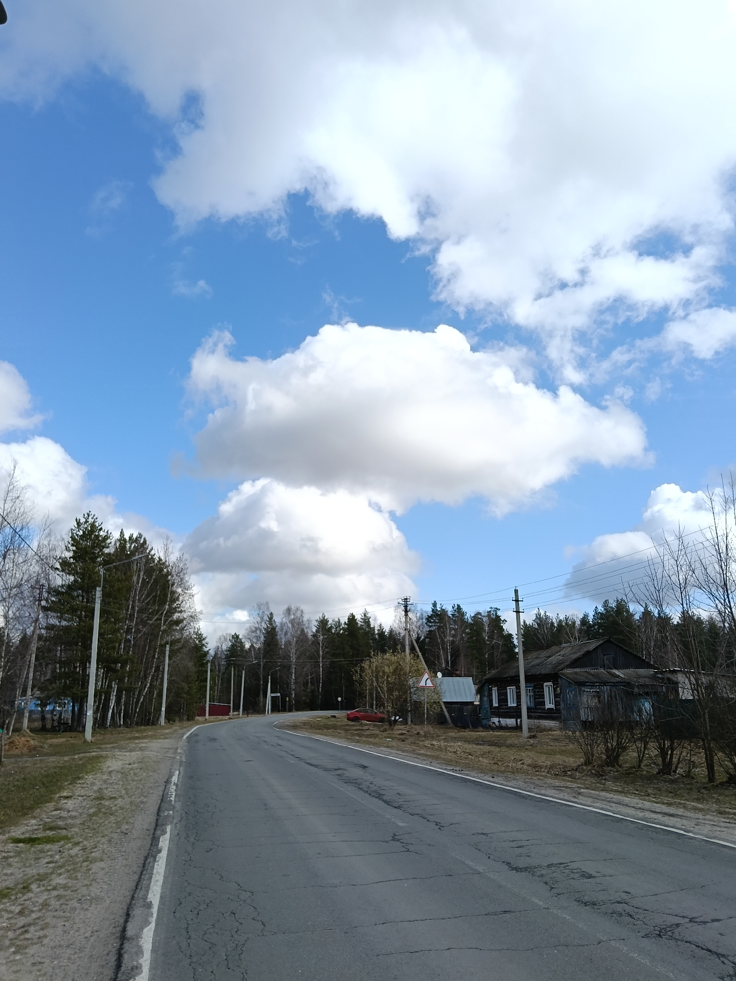 The image depicts a street scene in Красный Маяк, Russia, under a blue sky dotted with puffy white clouds. A gray asphalt road stretches into the distance, curving gently between lines of trees and power poles. The road surface shows signs of wear and cracking.
On the right side of the road, a dark, weathered wooden house with white-framed windows stands amidst dry grass and bare shrubs. In the background, a dense forest of coniferous trees is visible. On the left side of the road, there is a grassy shoulder and more trees, some with bare branches. A red structure is also visible on the left side of the road.