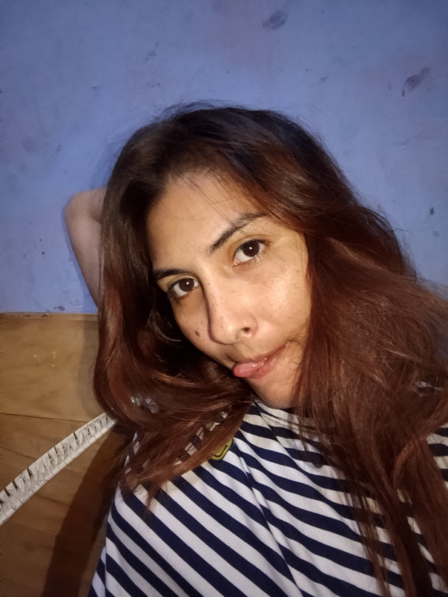 A young woman with long, brown hair and light skin poses for a selfie in an indoor setting. She is wearing a striped shirt with alternating white and dark blue lines. Her head is tilted slightly, and her tongue is playfully sticking out. Freckles are visible on her face. Her right arm is raised and resting behind her head. The background consists of a textured blue wall and a wooden surface behind her left arm. The location is Juan Griego, Venezuela.