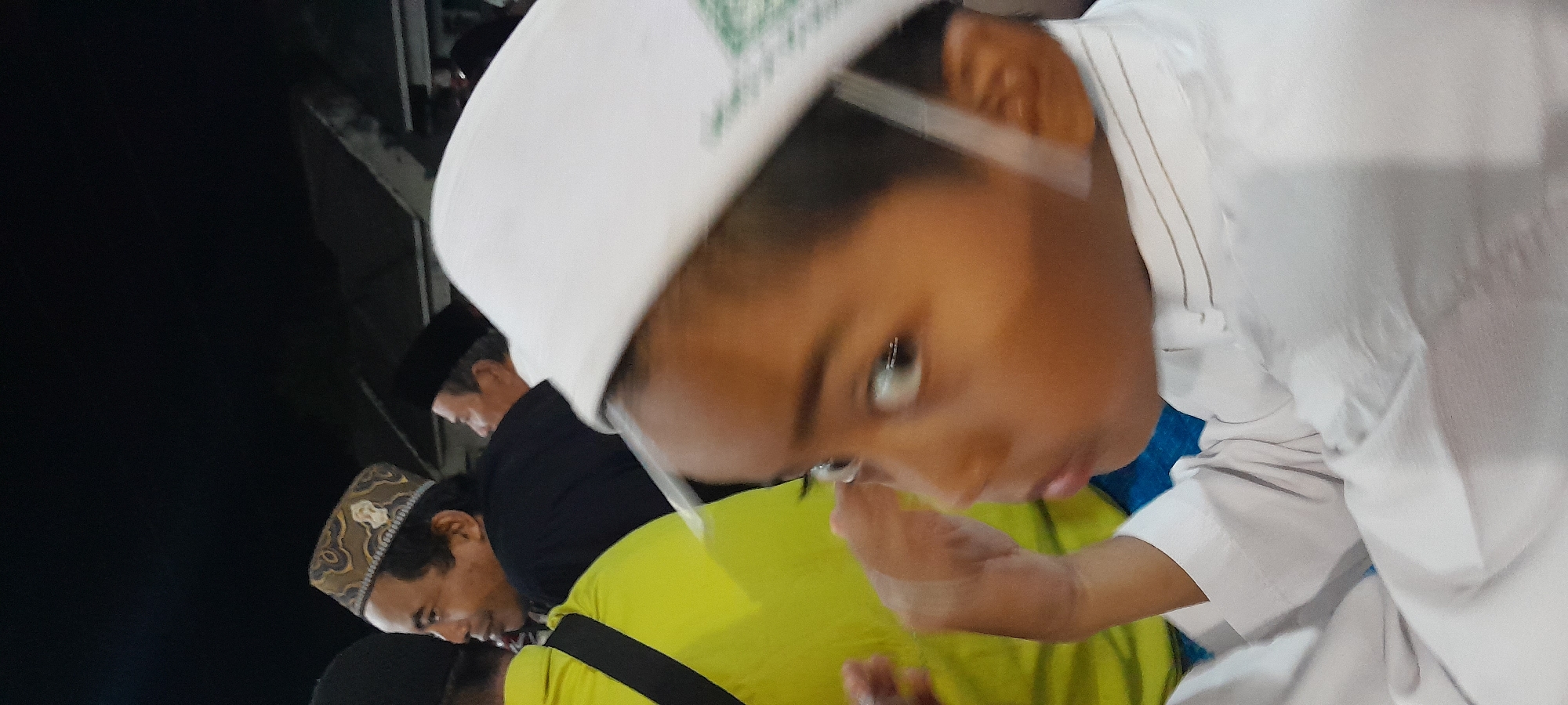 A close-up, side-profile shot captures a young boy in the foreground, wearing a white cap and white shirt with subtle pinstripes. His face is turned to the right, his eyes wide and reflective, with a light-colored band across his forehead. His right hand is raised to his face, obscuring part of his cheek. In the background, out of focus, are several adults. One man to the left wears a yellow shirt and another to his right wears a black shirt and a patterned cap. The setting appears to be an outdoor gathering at night, possibly in Bendungan, Indonesia, with hints of stadium seating or elevated structures in the far background, illuminated by artificial lights. The overall impression is one of a candid moment during a community event.